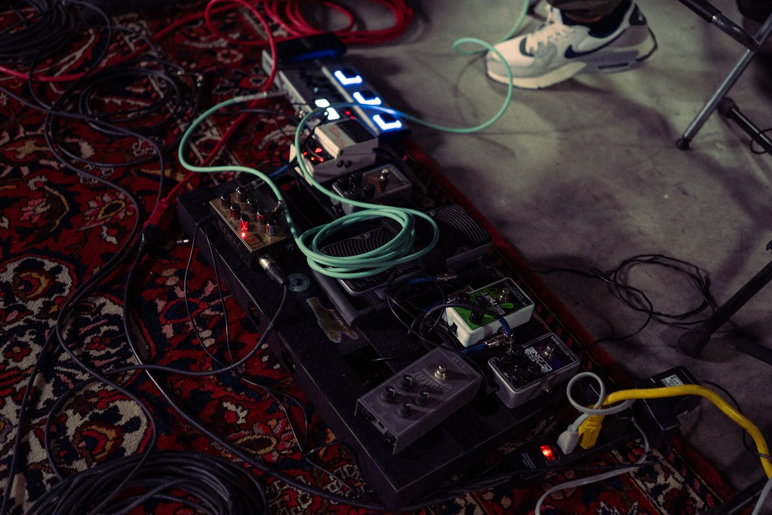 A pedalboard with multiple guitar effects pedals connected by various cables, placed on a patterned rug.