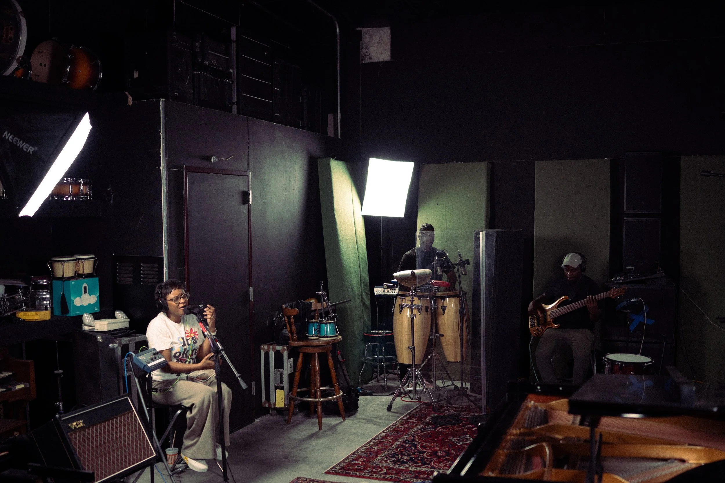 A recording studio with three musicians practicing: a woman singing into a microphone, a person playing conga drums, and a person playing bass guitar. Various musical equipment and instruments are visible, with black walls, some lighting, and a red p