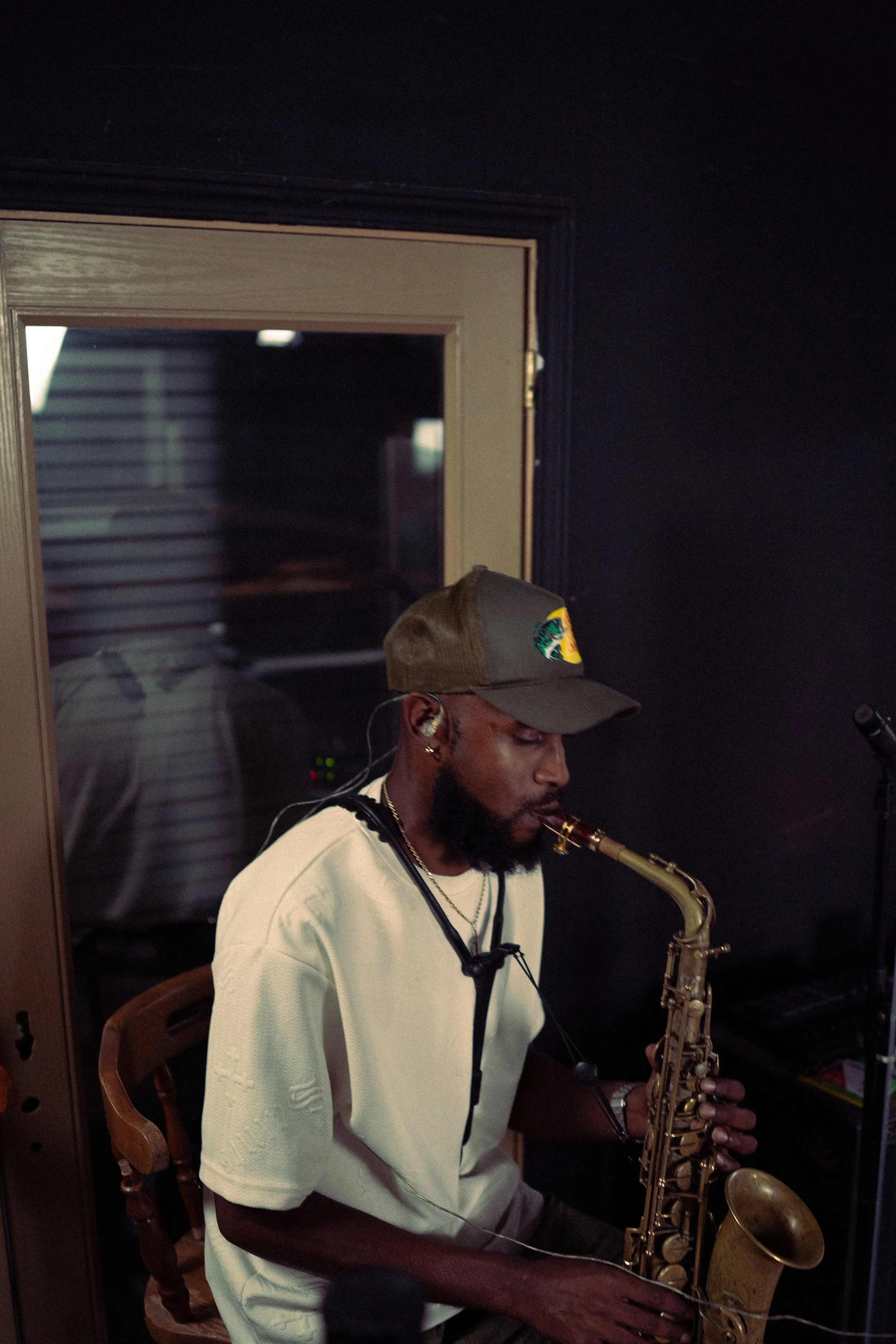 A man playing a saxophone in a dimly lit room, wearing a baseball cap and casual clothing.