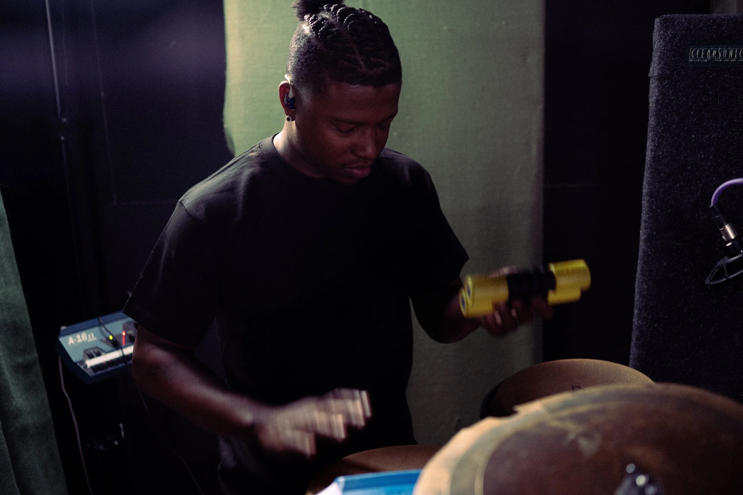 A person in a dark t-shirt in a recording studio holding a yellow and black bottle, surrounded by audio equipment and a percussion instrument.