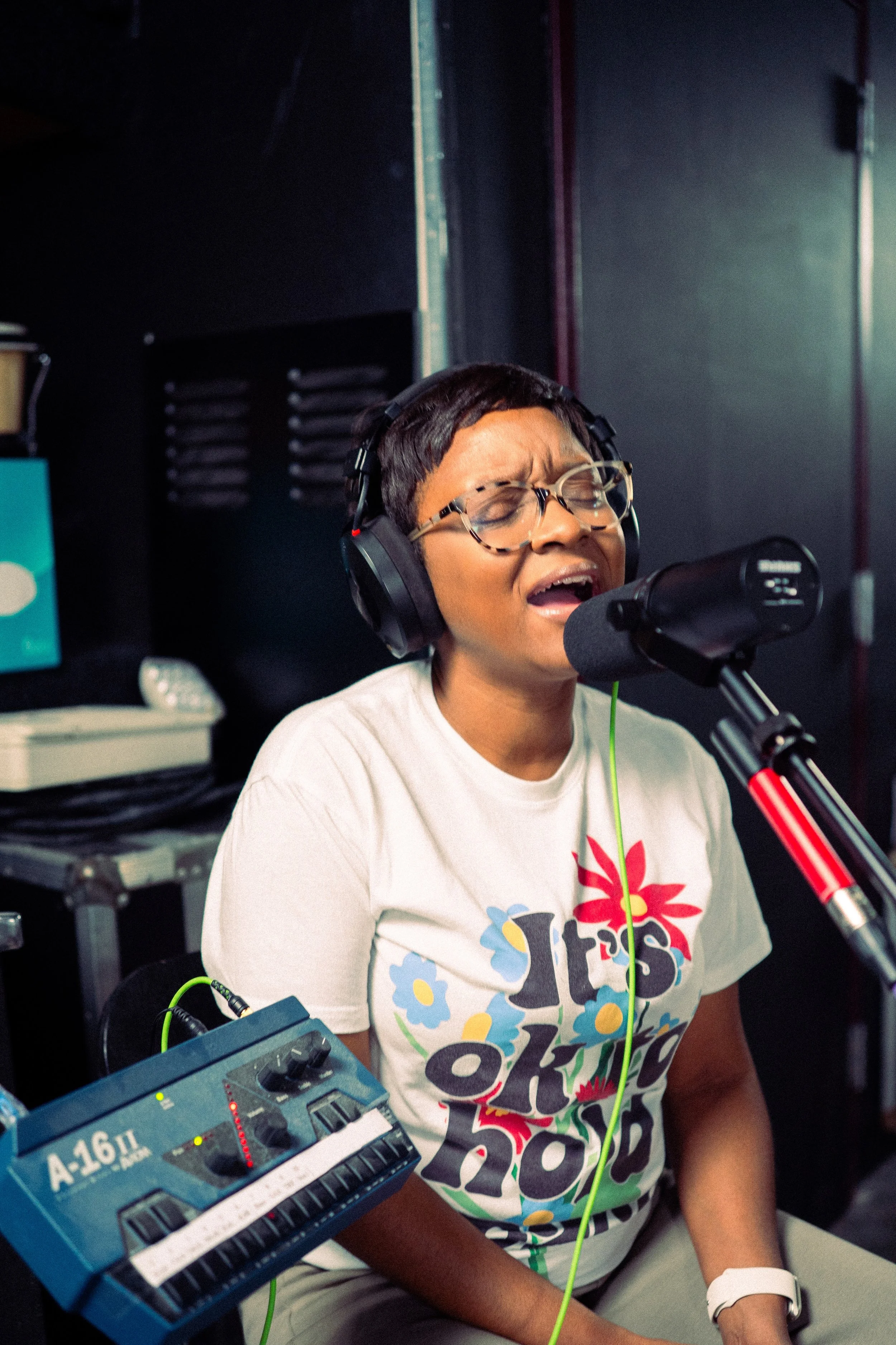 A woman in glasses and a white T-shirt singing into a microphone in a recording studio.