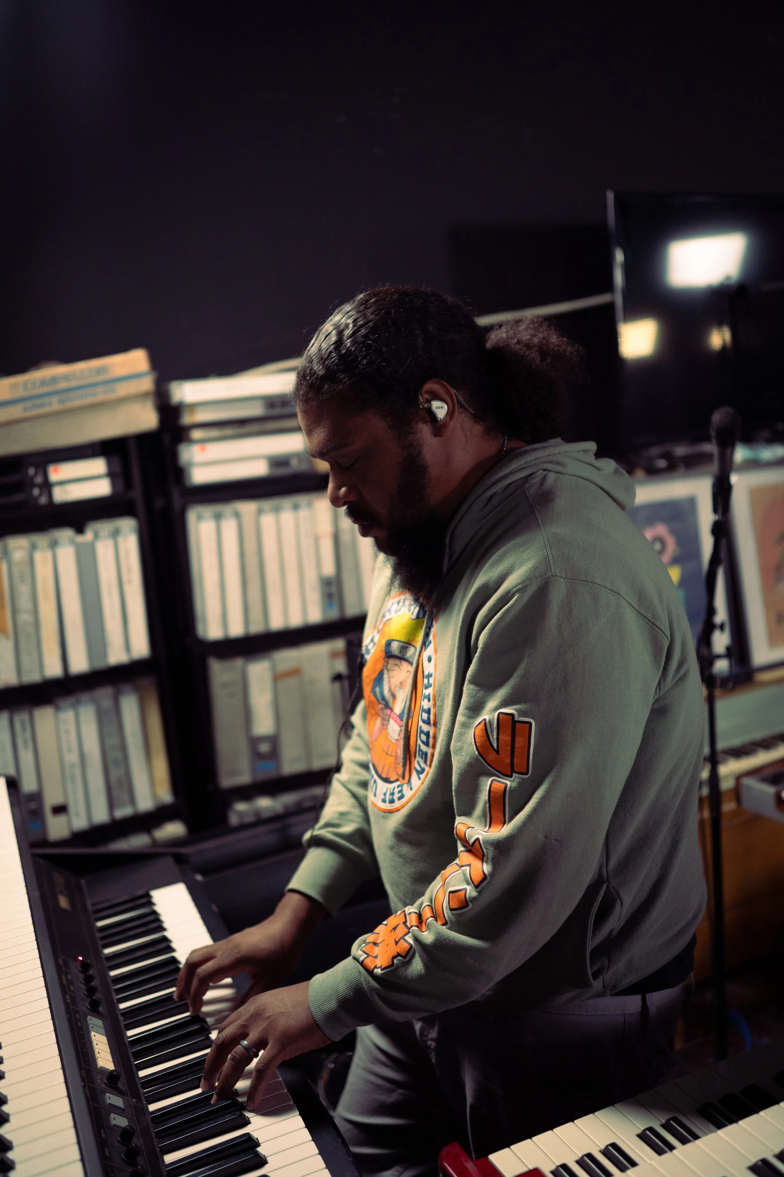 A man playing keyboard in a music studio with shelves of books and a television in the background.