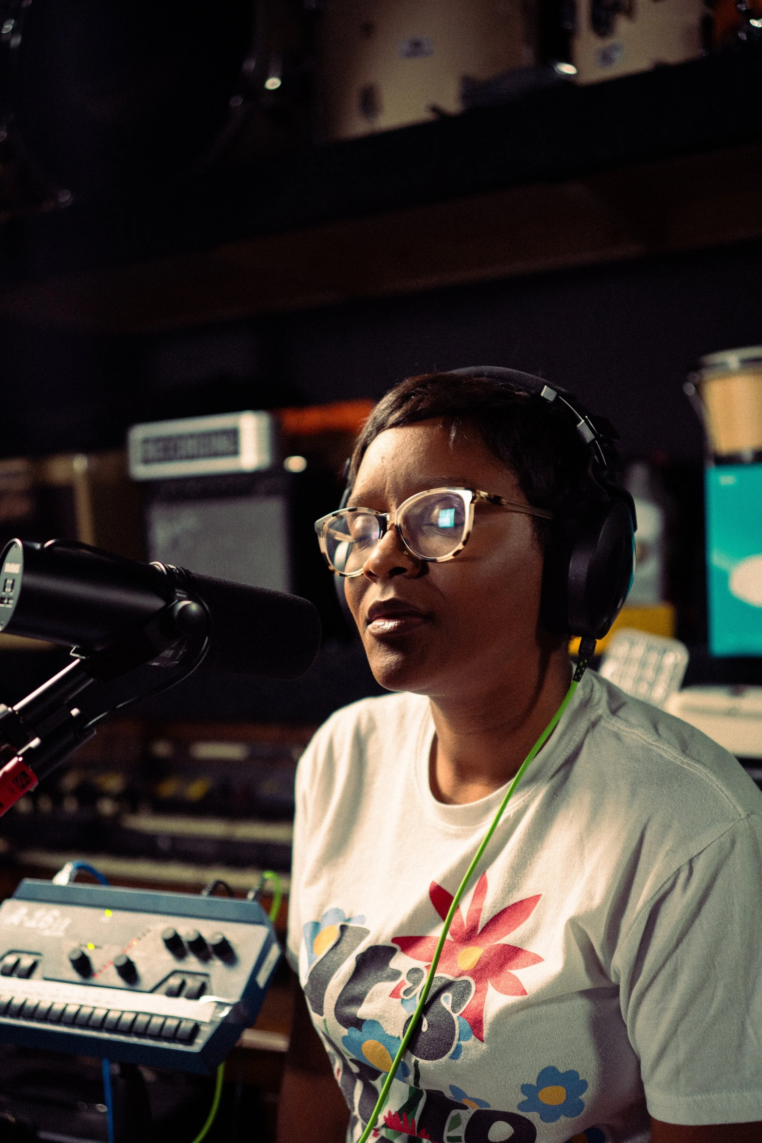 A woman with short hair, glasses, and headphones is speaking into a microphone in a recording studio, surrounded by audio equipment and monitors.