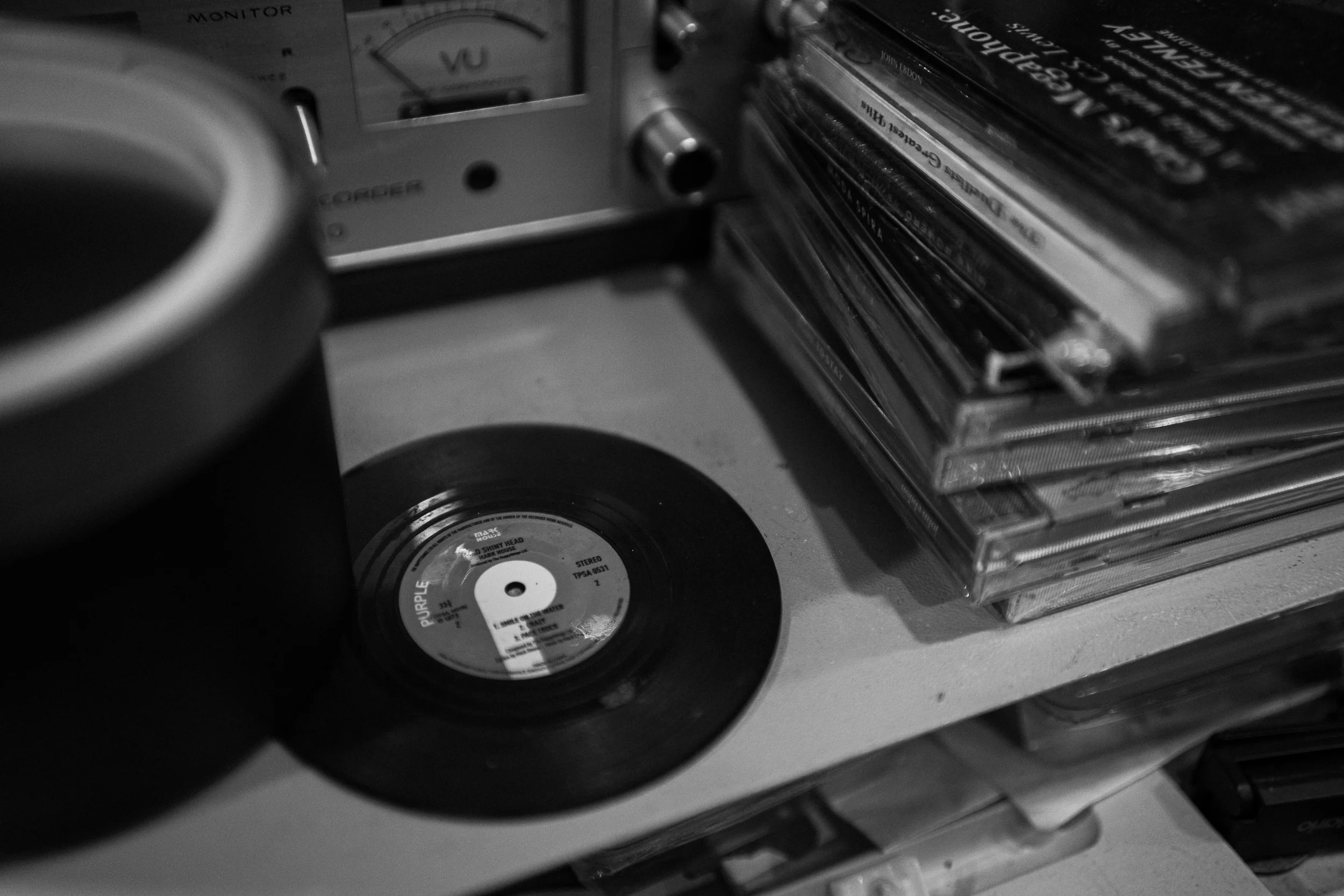 A black and white photo of vintage music items, including a small vinyl record, a stack of CD cases, a radio, and a coffee mug on a table.