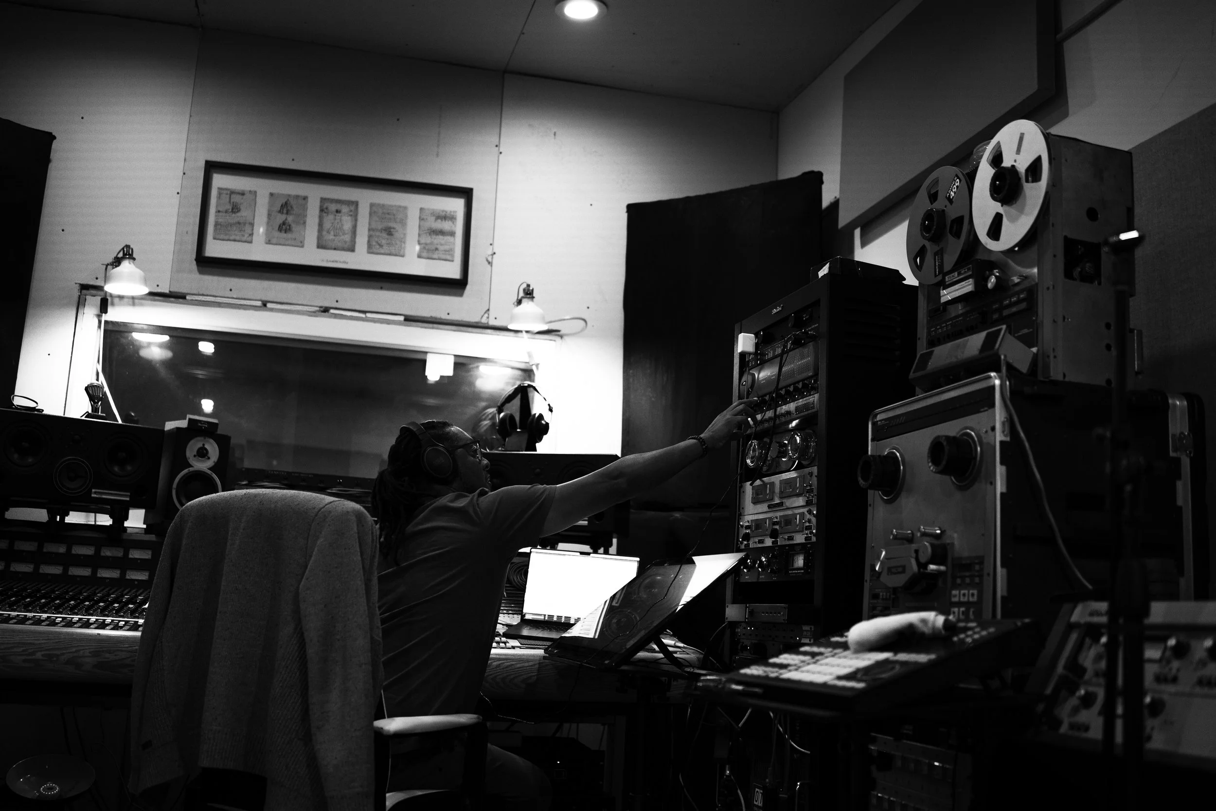 A person in a recording studio working with audio equipment, wearing headphones and reaching for controls among racks of audio gear, with a mirrored window reflecting part of the room.