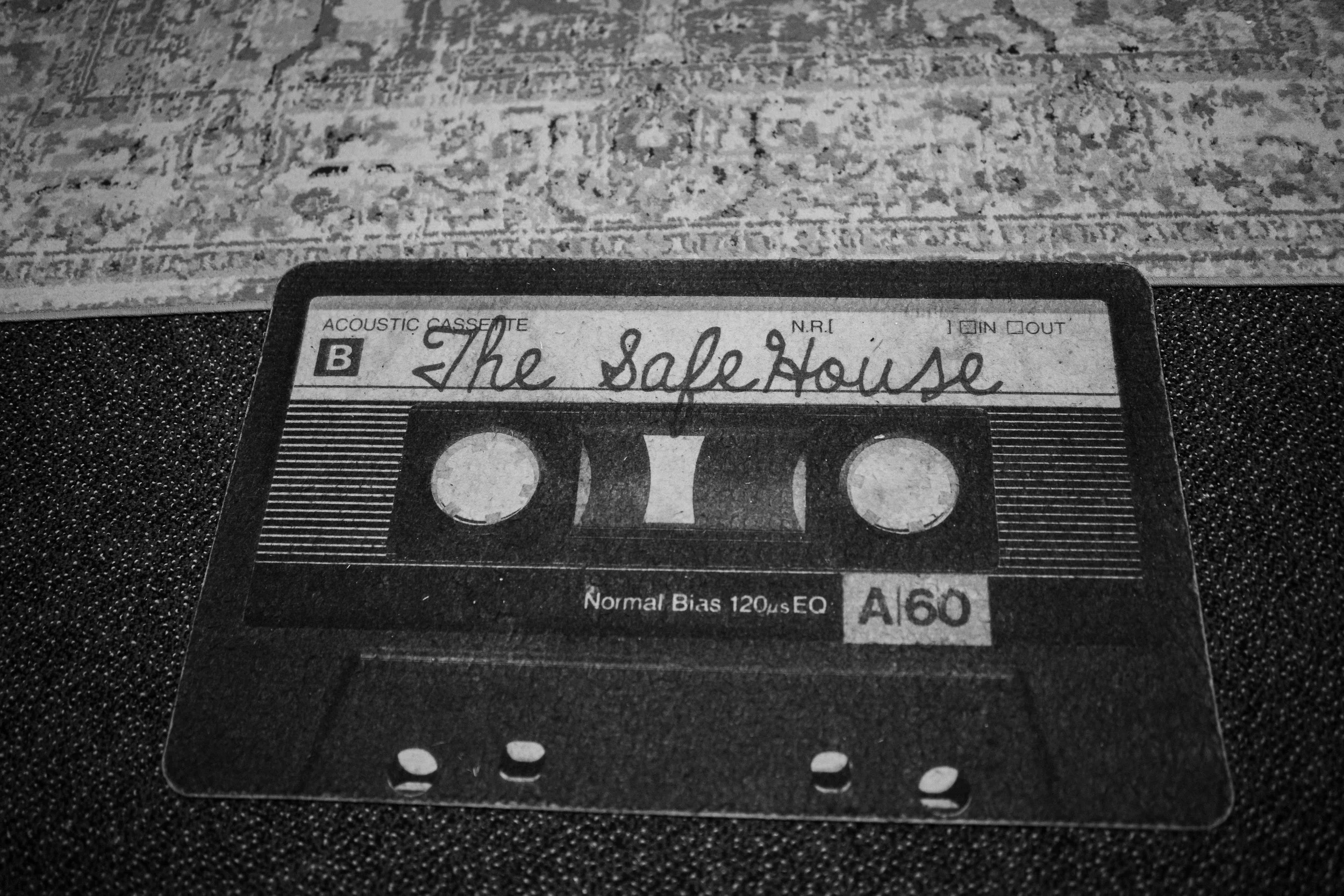 A black and white photograph of a cassette tape labeled "The Safe House" resting on a textured surface with a patterned carpet in the background.
