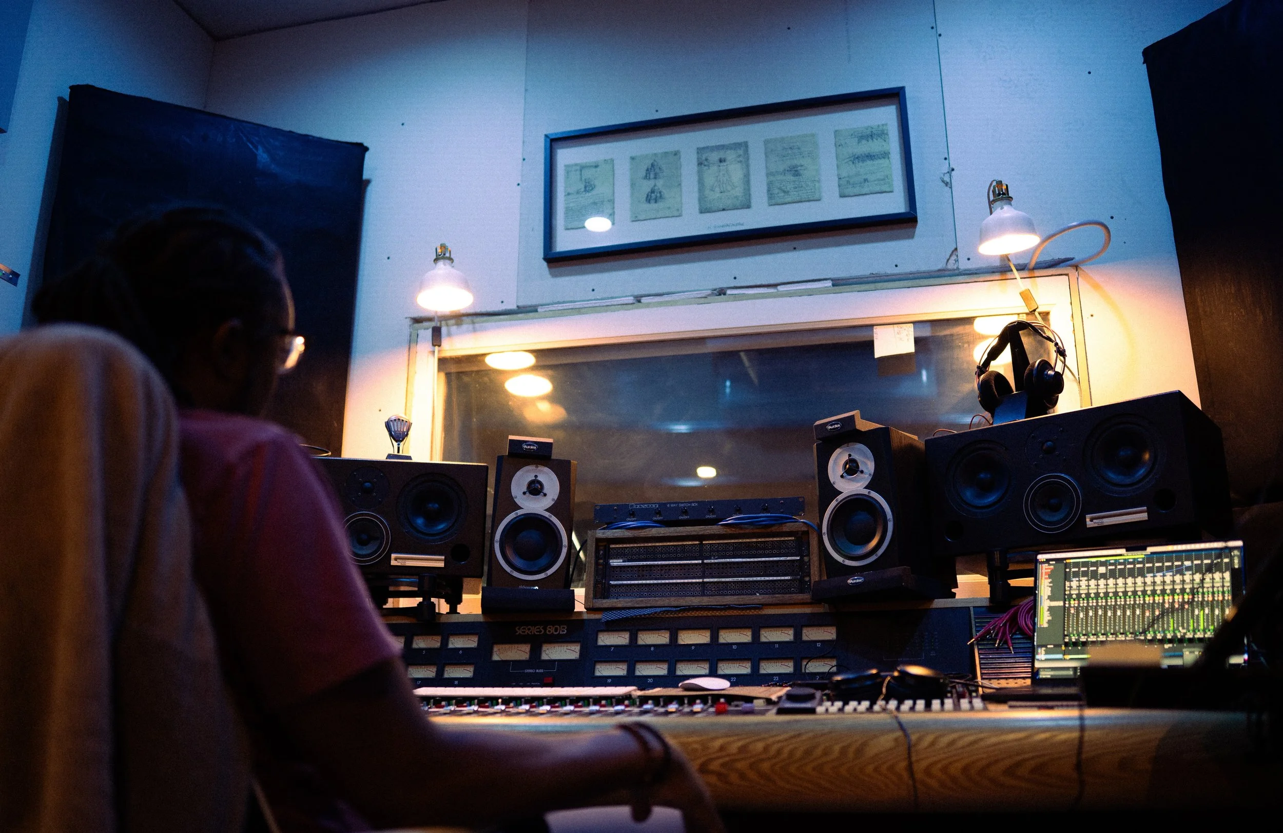 Recording studio with person working at mixing console, professional speakers, headphones, and sound equipment. dim lighting, framed artwork above window.