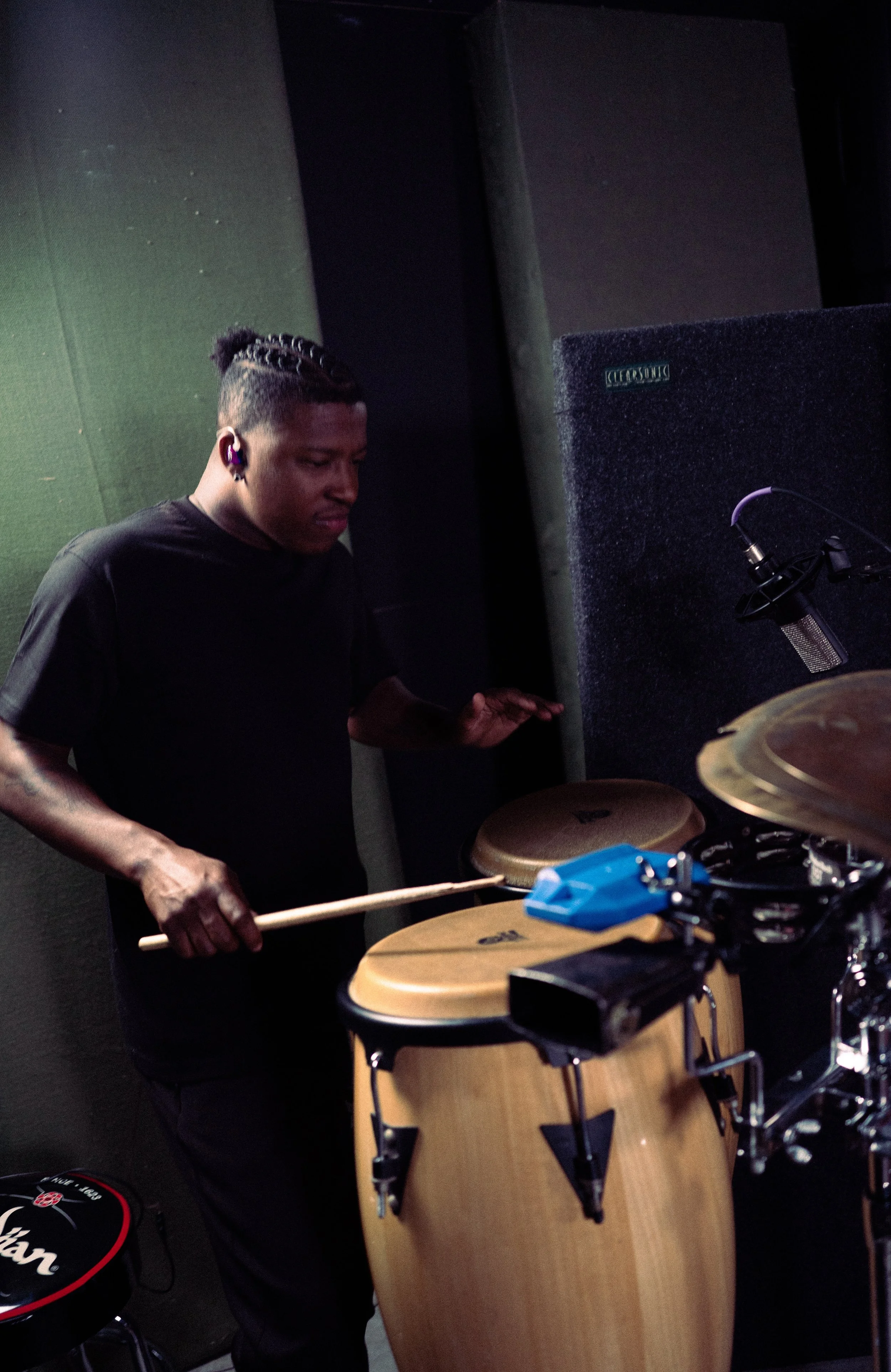 A young man with braided hair playing conga drums in a music studio, wearing headphones and focused on his performance.