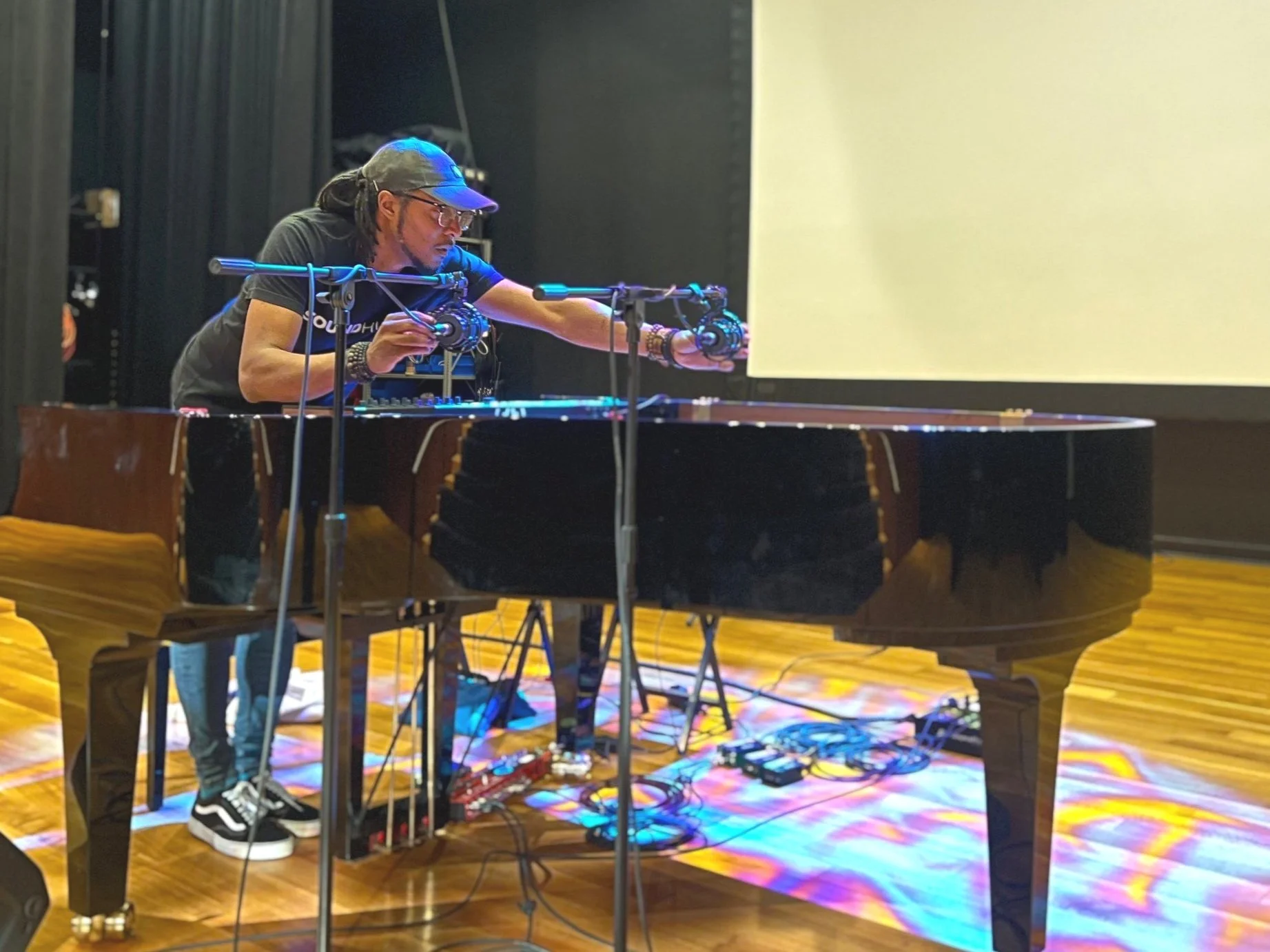 Person with glasses and cap adjusting DJ equipment on a grand piano in a dimly lit room.