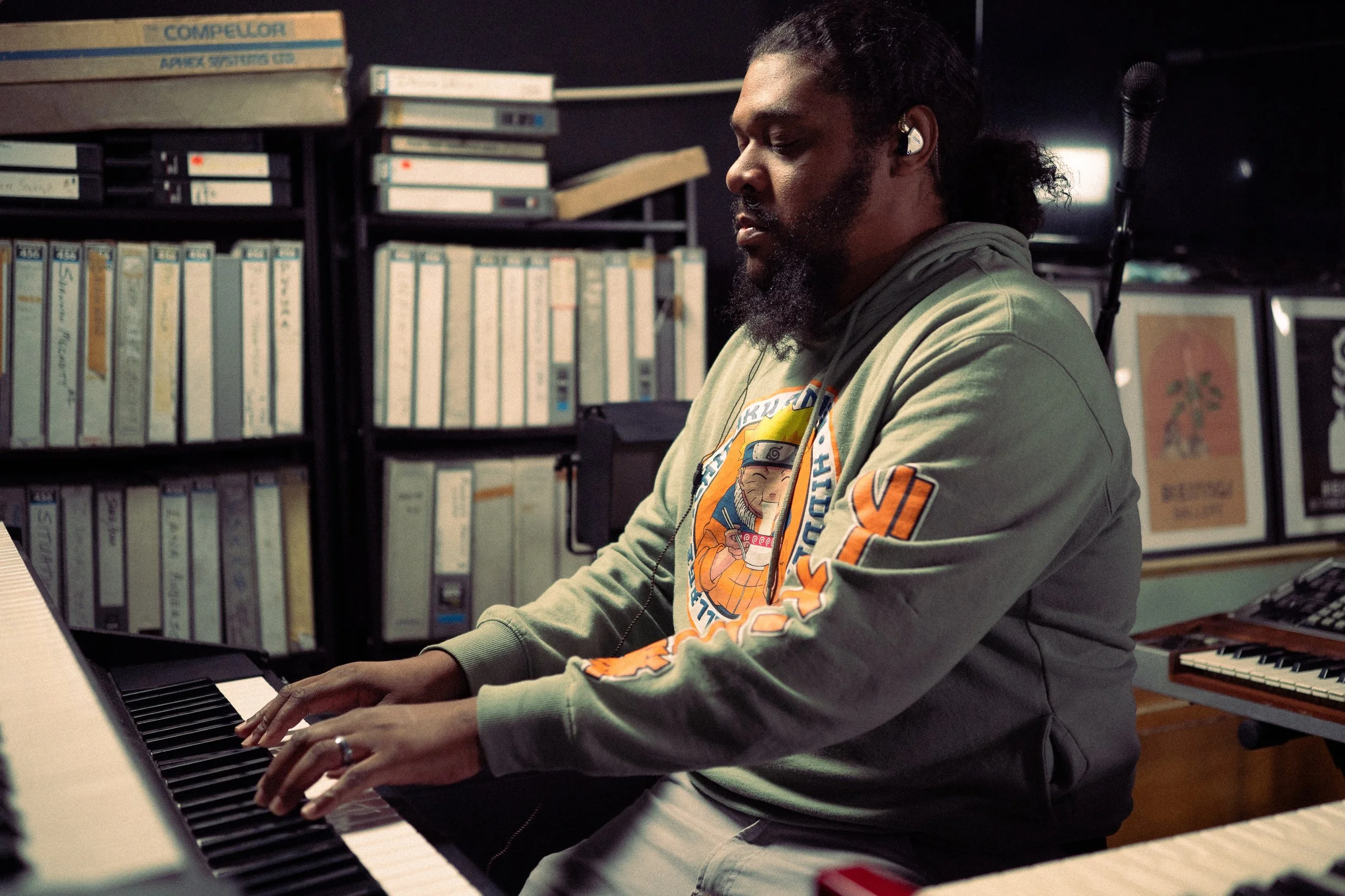 A man with long hair and a beard wearing headphones plays a keyboard in a room filled with music equipment and shelves with books and framed pictures.