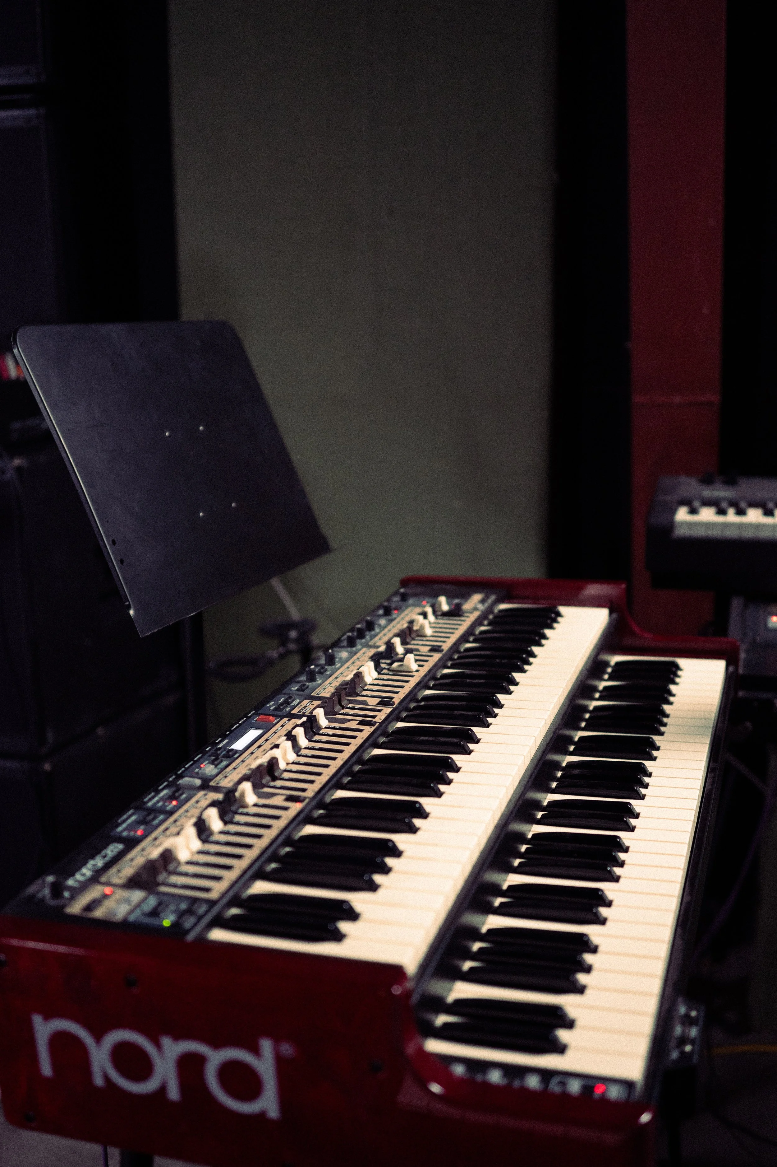 A red Nord electronic keyboard on a stand with a sheet music stand nearby, located in a music studio.