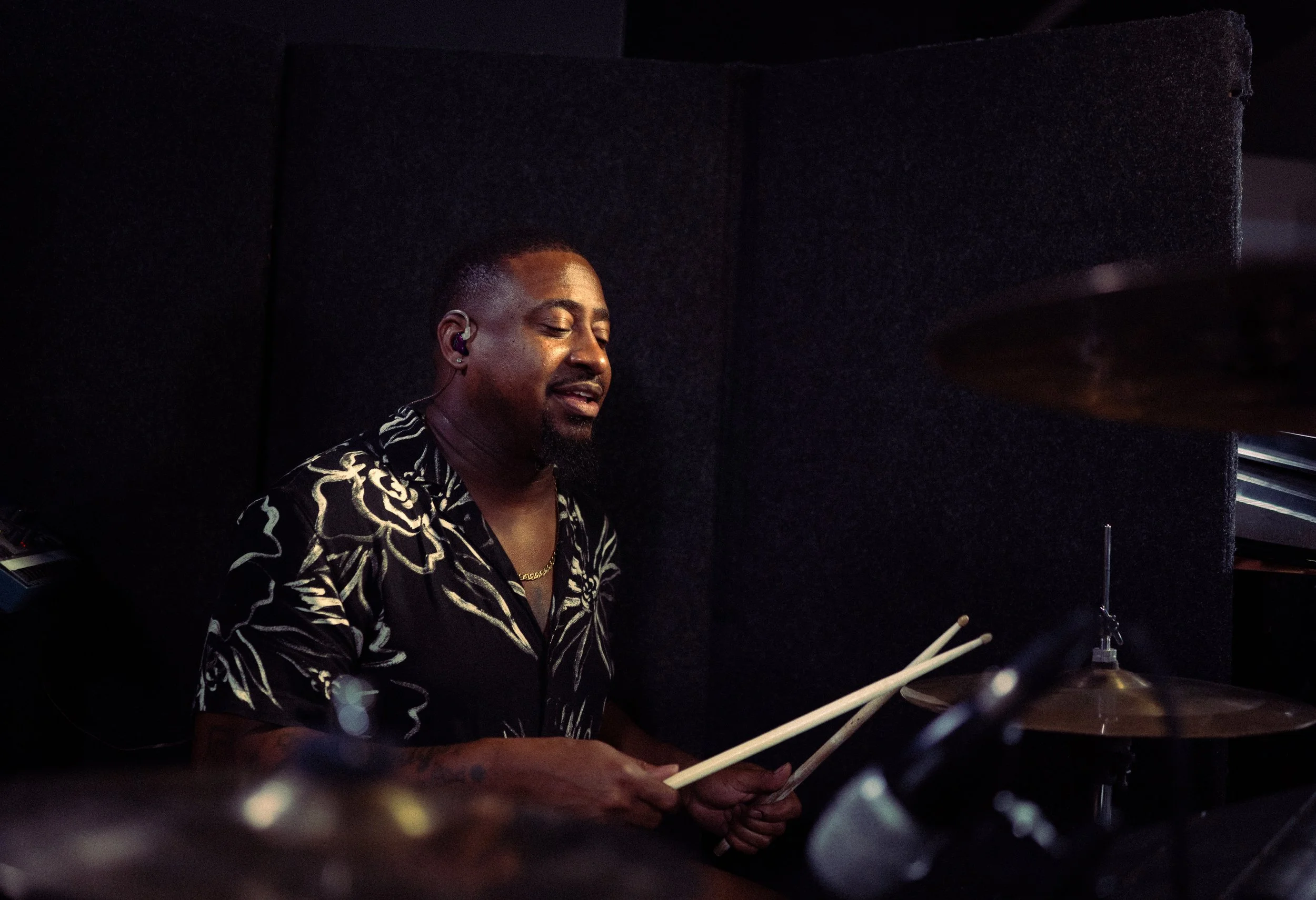 Man playing drums in a music studio, wearing a black shirt with a floral pattern, with closed eyes and holding drumsticks.