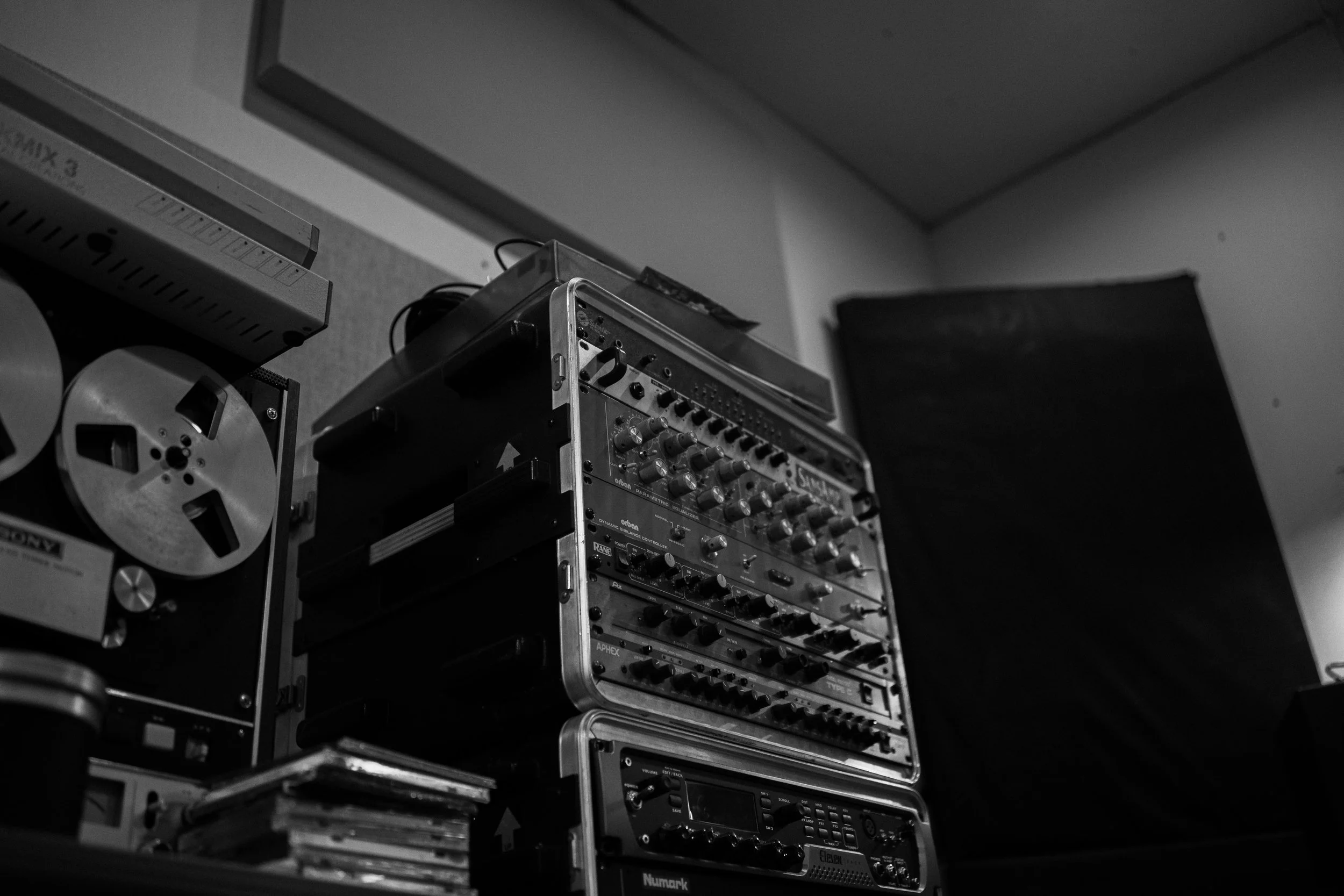 Black and white image of audio equipment, including a reel-to-reel tape recorder, audio mixer, and audio receiver, stacked on top of each other, in a recording studio.