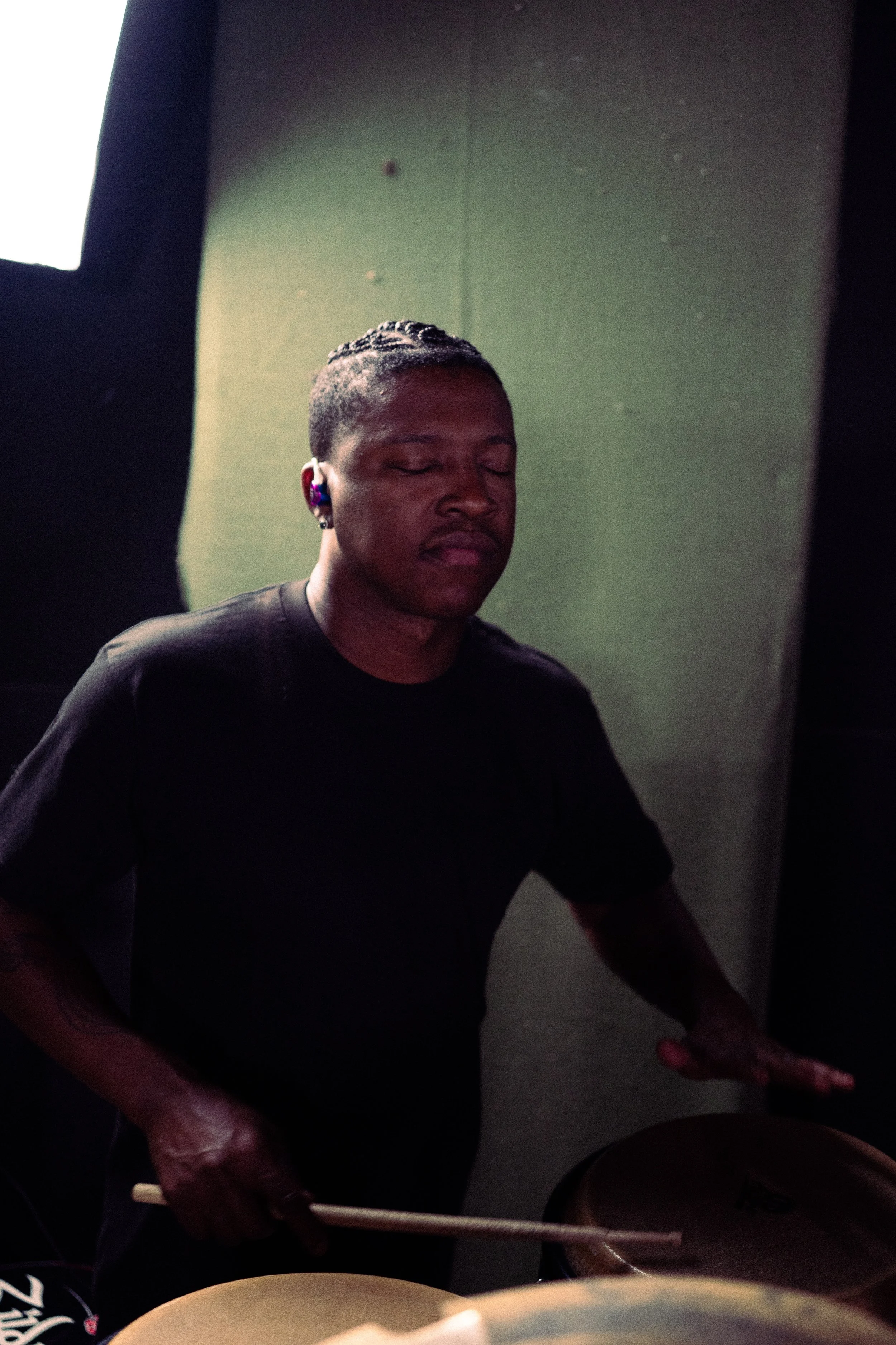 A person with braided hair and wearing a black shirt is playing drums, eyes closed, with a focused expression. They are in a dimly lit room with a greenish wall background.