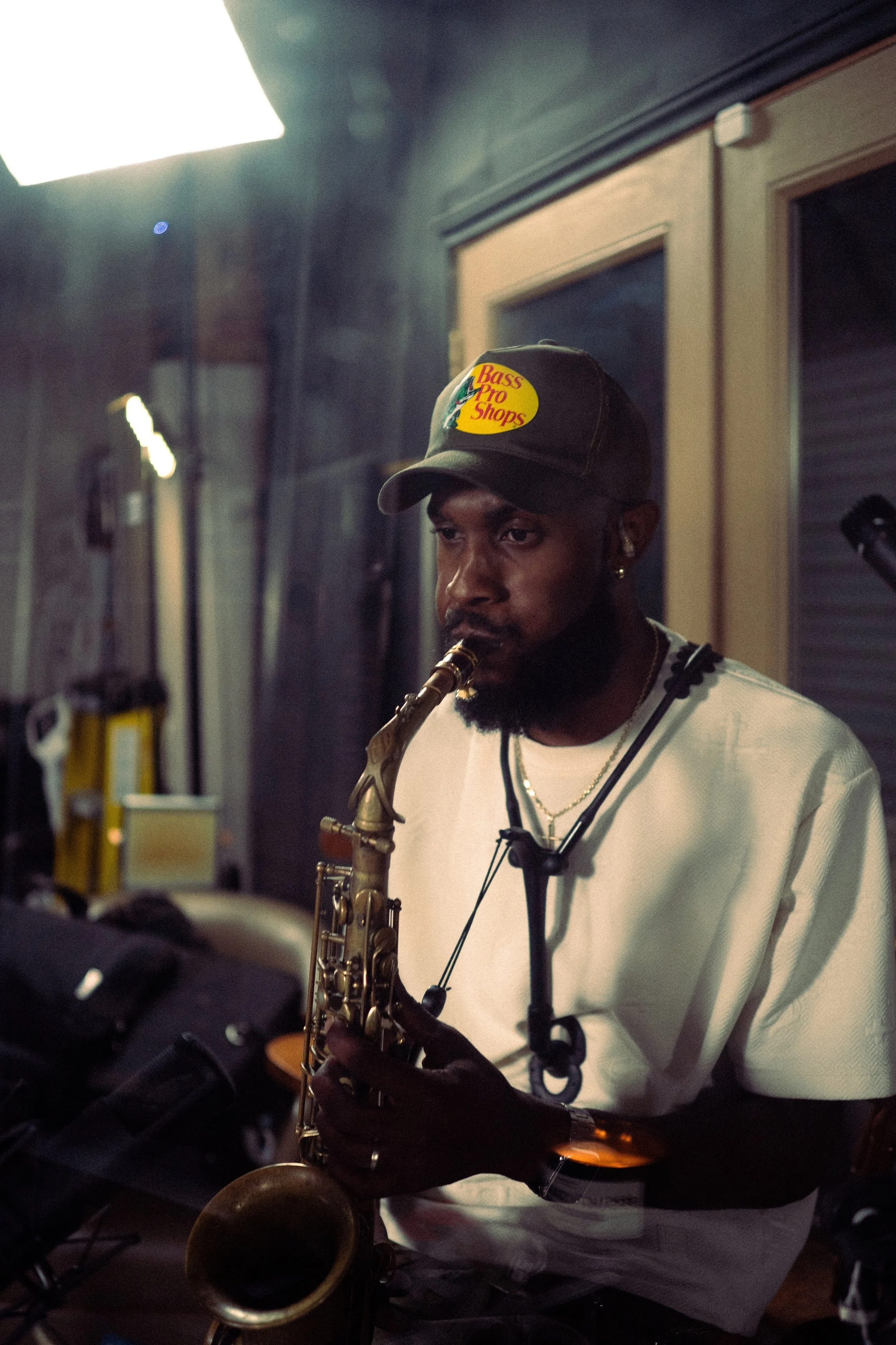 A man wearing a Bass Pro Shops cap playing a saxophone indoors, with musical equipment and equipment in the background.