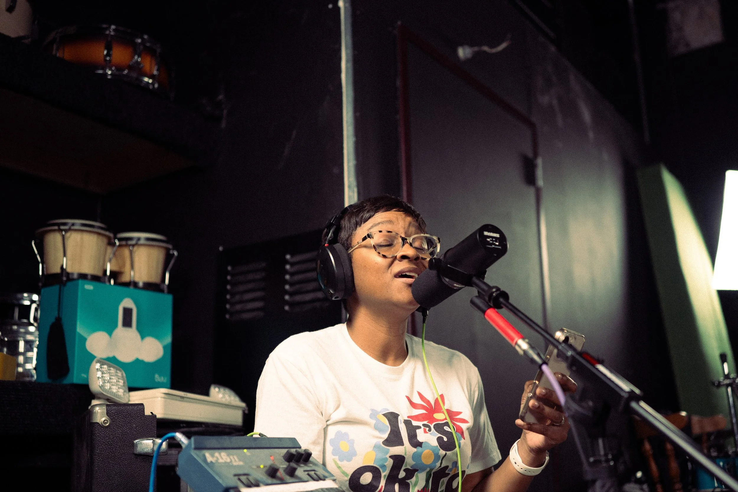 A person singing into a microphone in a music studio, wearing headphones and glasses, holding a phone, with musical instruments and equipment in the background.