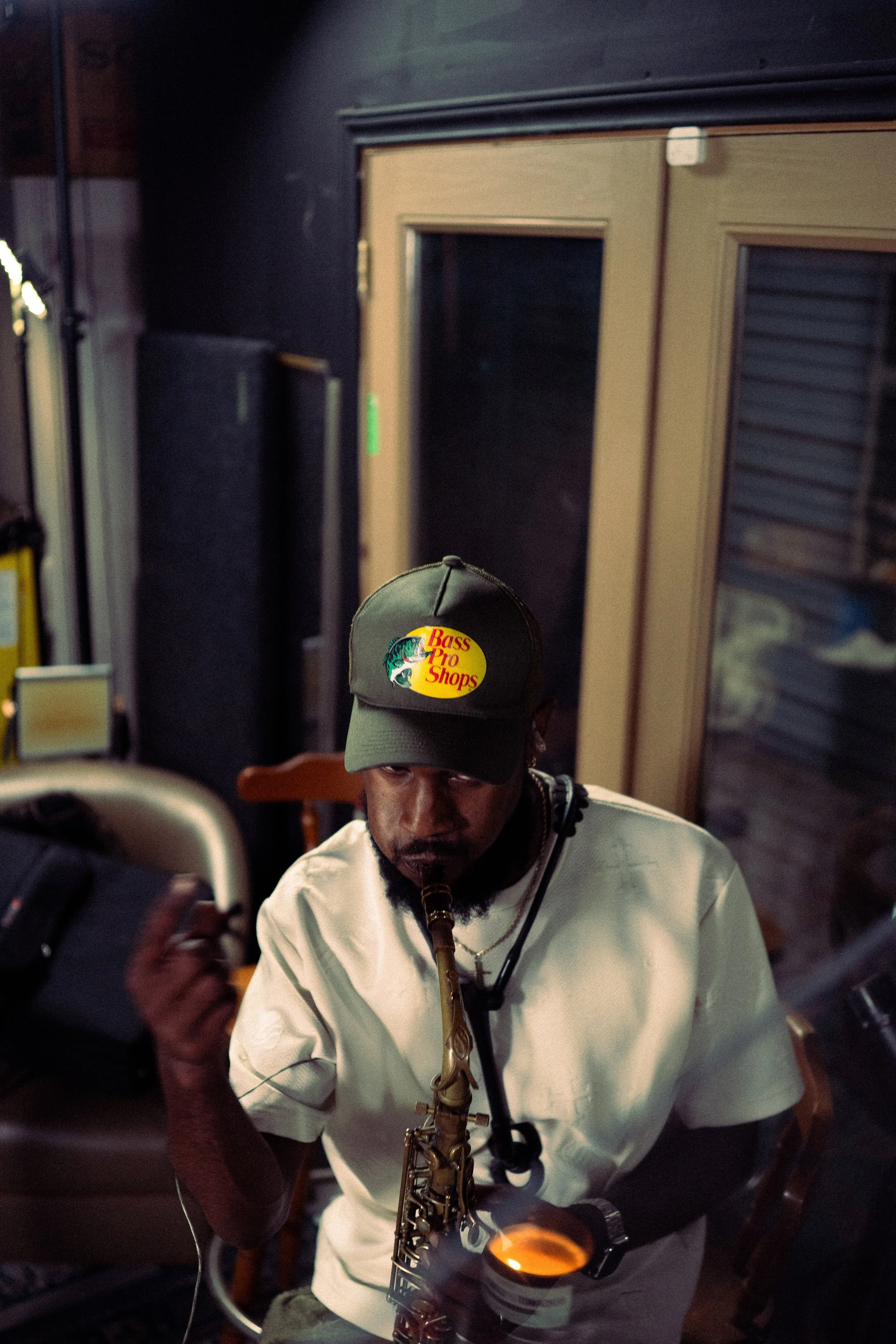A man wearing a Bass Pro Shops cap is playing a saxophone while drinking a beverage in a dimly lit room.