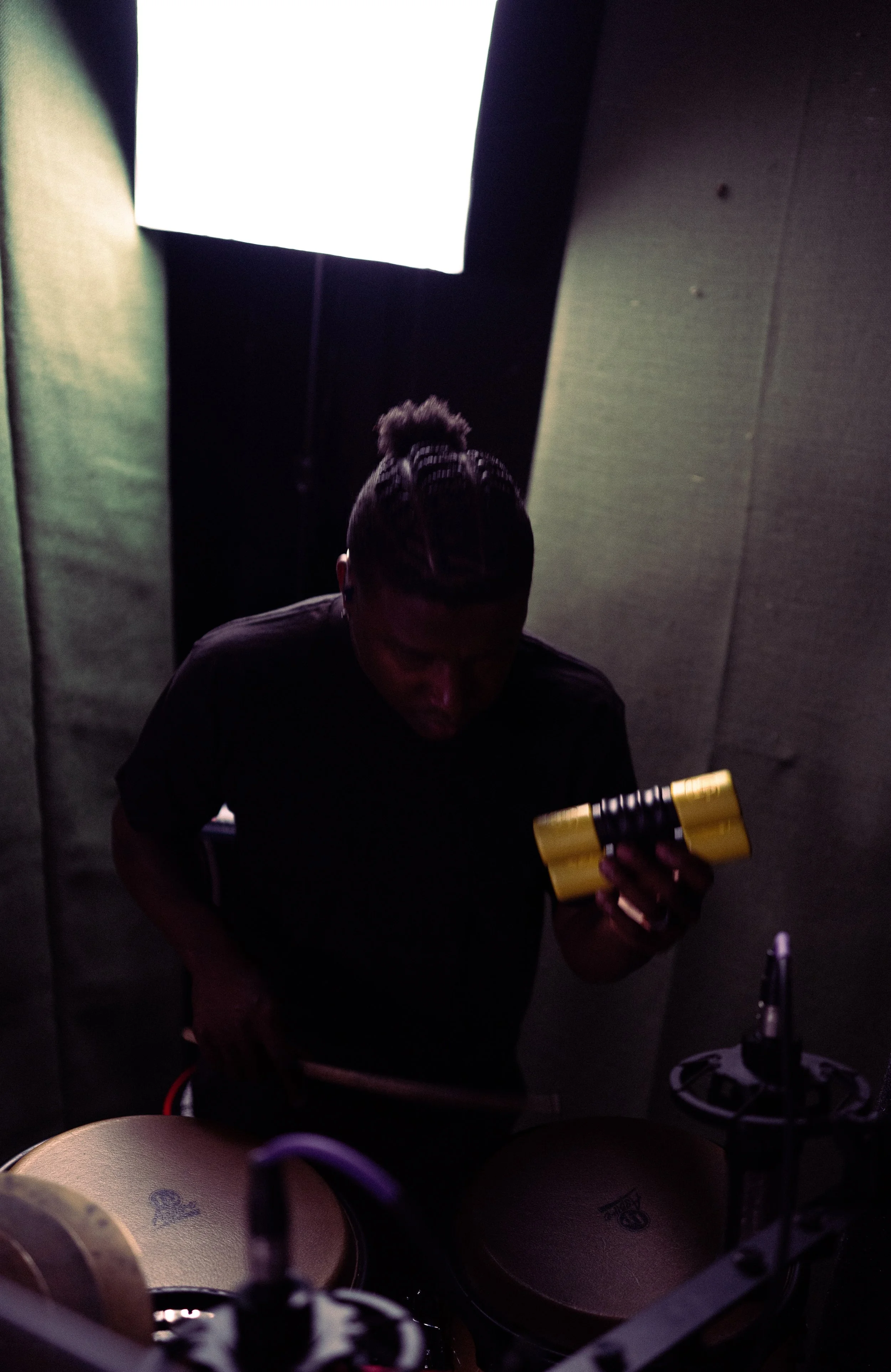 Person with braided hair looking at their phone in a dark room with percussion instruments and bright light in the background.