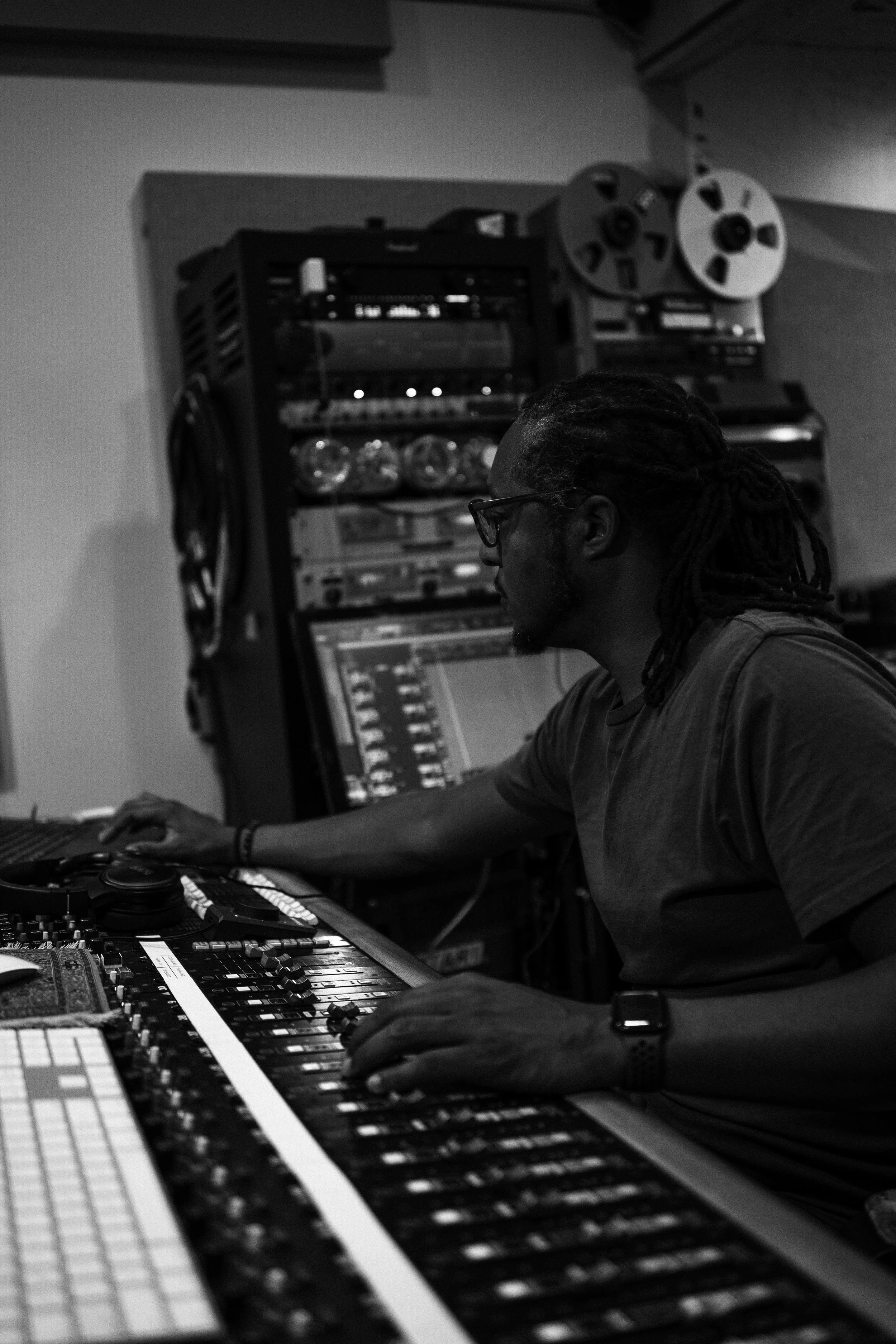 A man with dreadlocks and glasses working on a music mixing console in a recording studio, with reel-to-reel tape machines and audio equipment in the background.