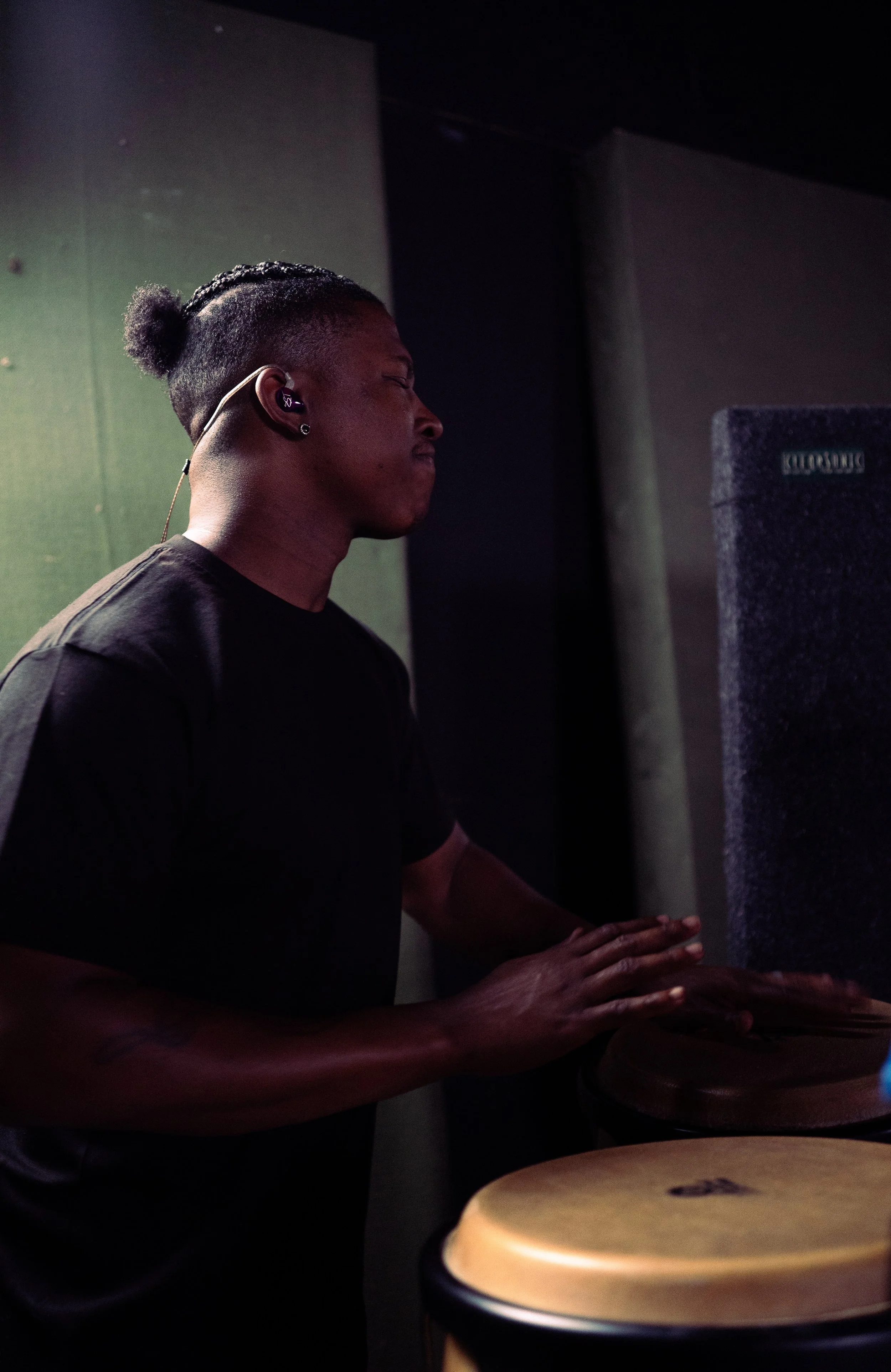 A person playing conga drums in a dimly lit room, wearing a black shirt and in-ear monitors.