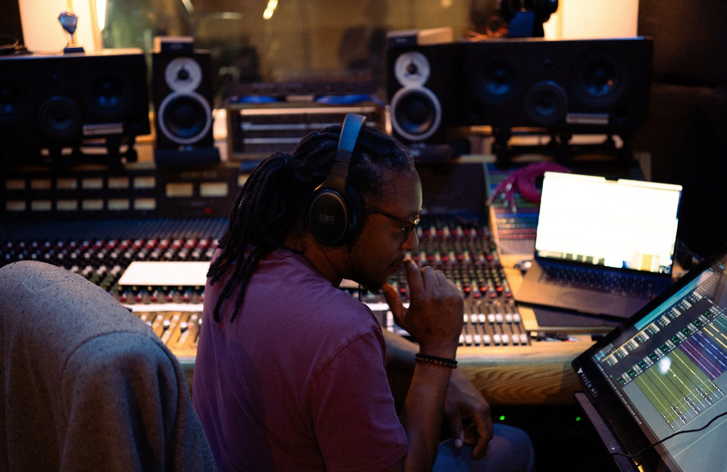 A person with long dreadlocks wearing headphones and glasses, sitting in front of a large audio mixing console, working on music production in a studio with monitors and a laptop.