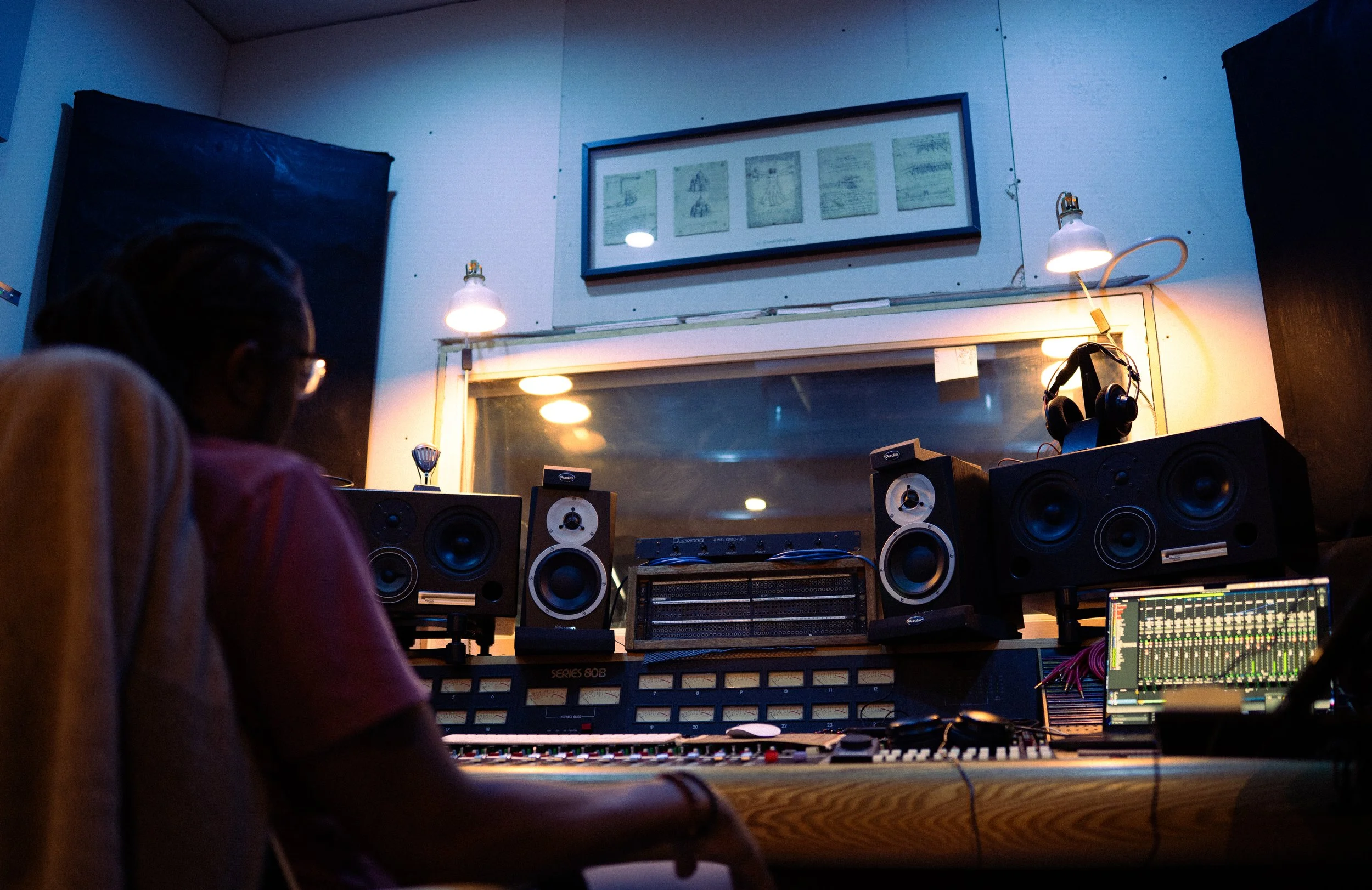 Inside a recording studio with a person working on audio equipment, studio monitors, a computer with sound mixing software, and framed artwork on the wall.