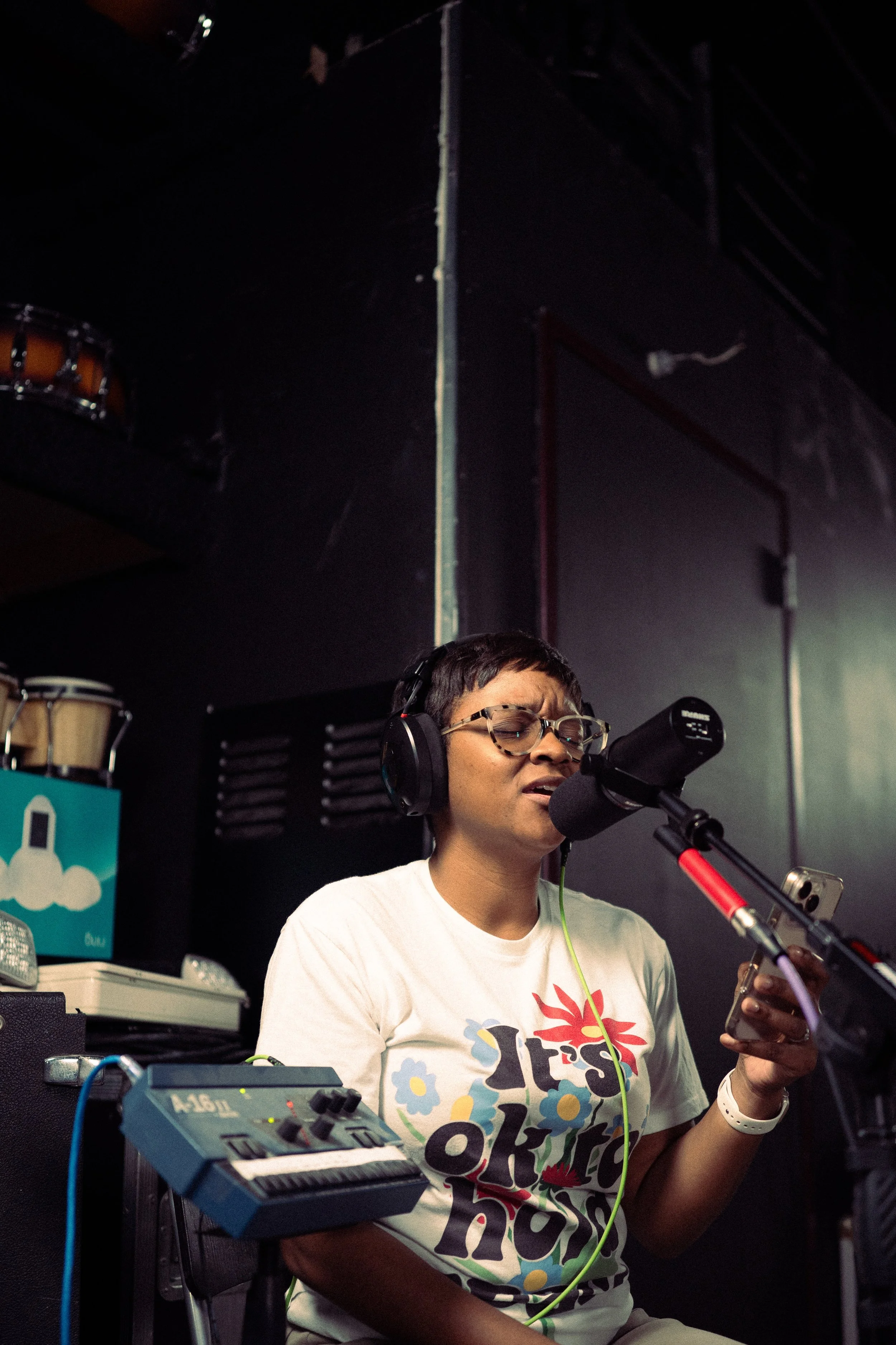 A person with short hair, glasses, and a white T-shirt singing into a microphone in a recording studio, wearing headphones, holding a phone in one hand.