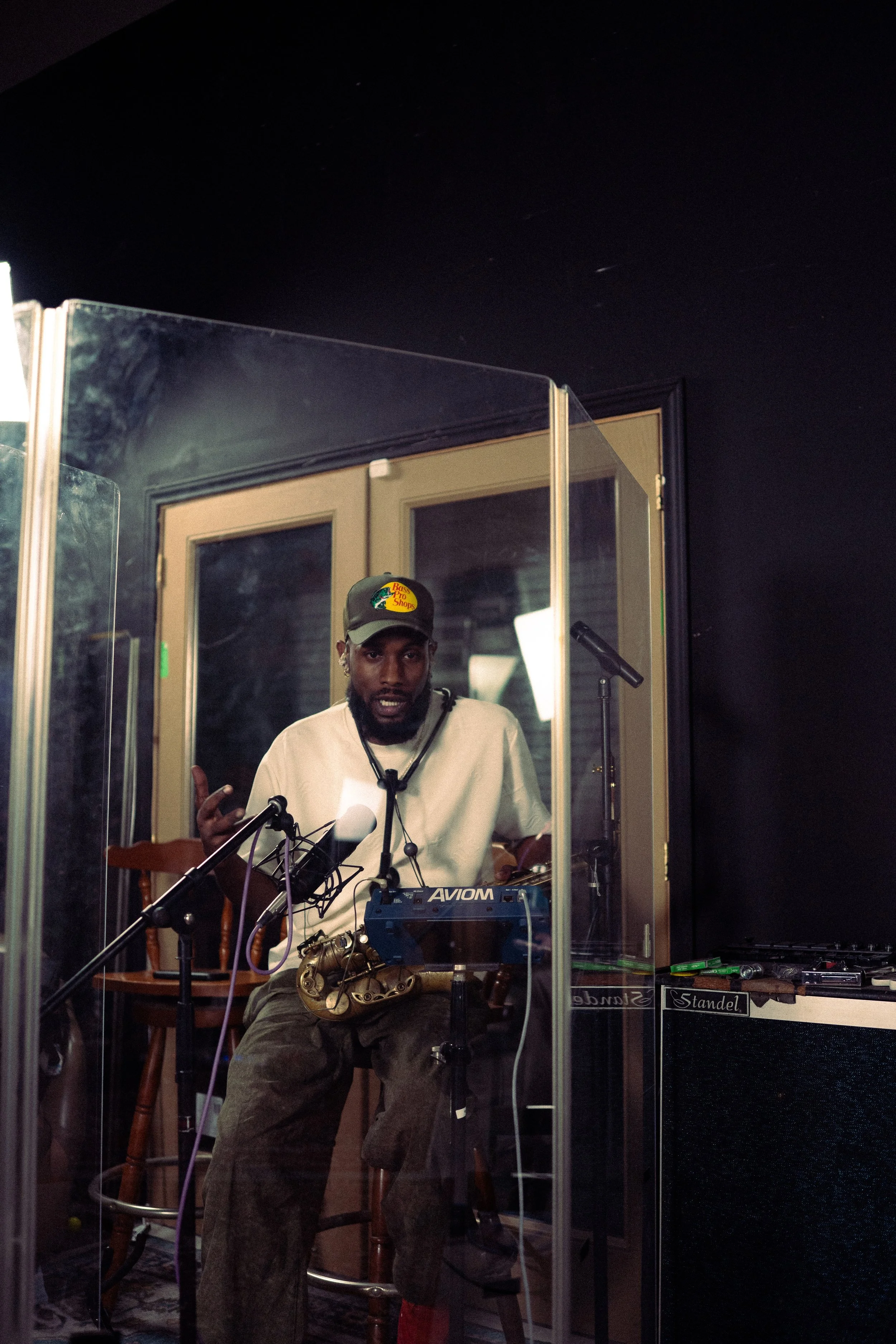 A man in a baseball cap and a white t-shirt sitting behind a glass partition in a recording studio, with a microphone, musical instrument, and sound equipment.