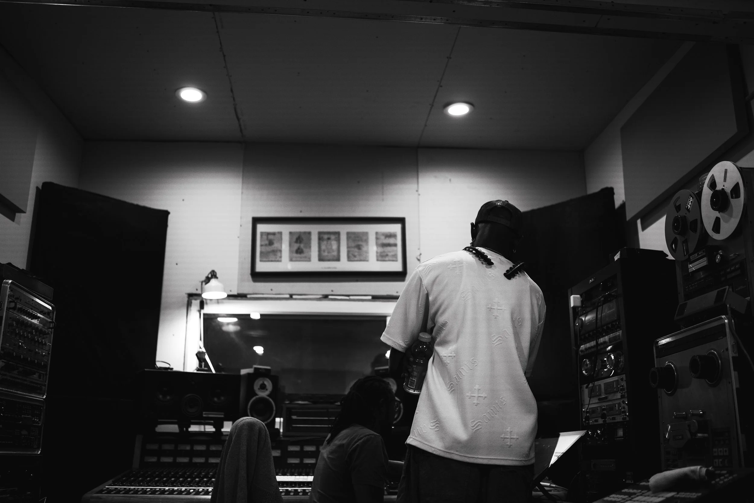 Black and white photo inside a music recording studio with two people working. One person standing holding a water bottle, the other sitting at a mixing console.