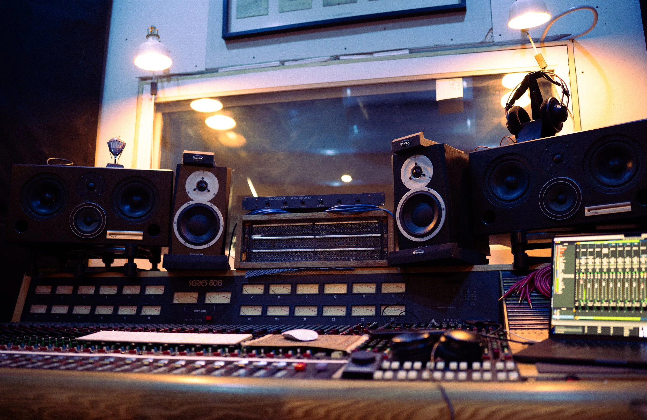 A professional recording studio control room with mixing console, studio monitors, a laptop, and audio equipment, illuminated by overhead and side lights, and seen through a large window.