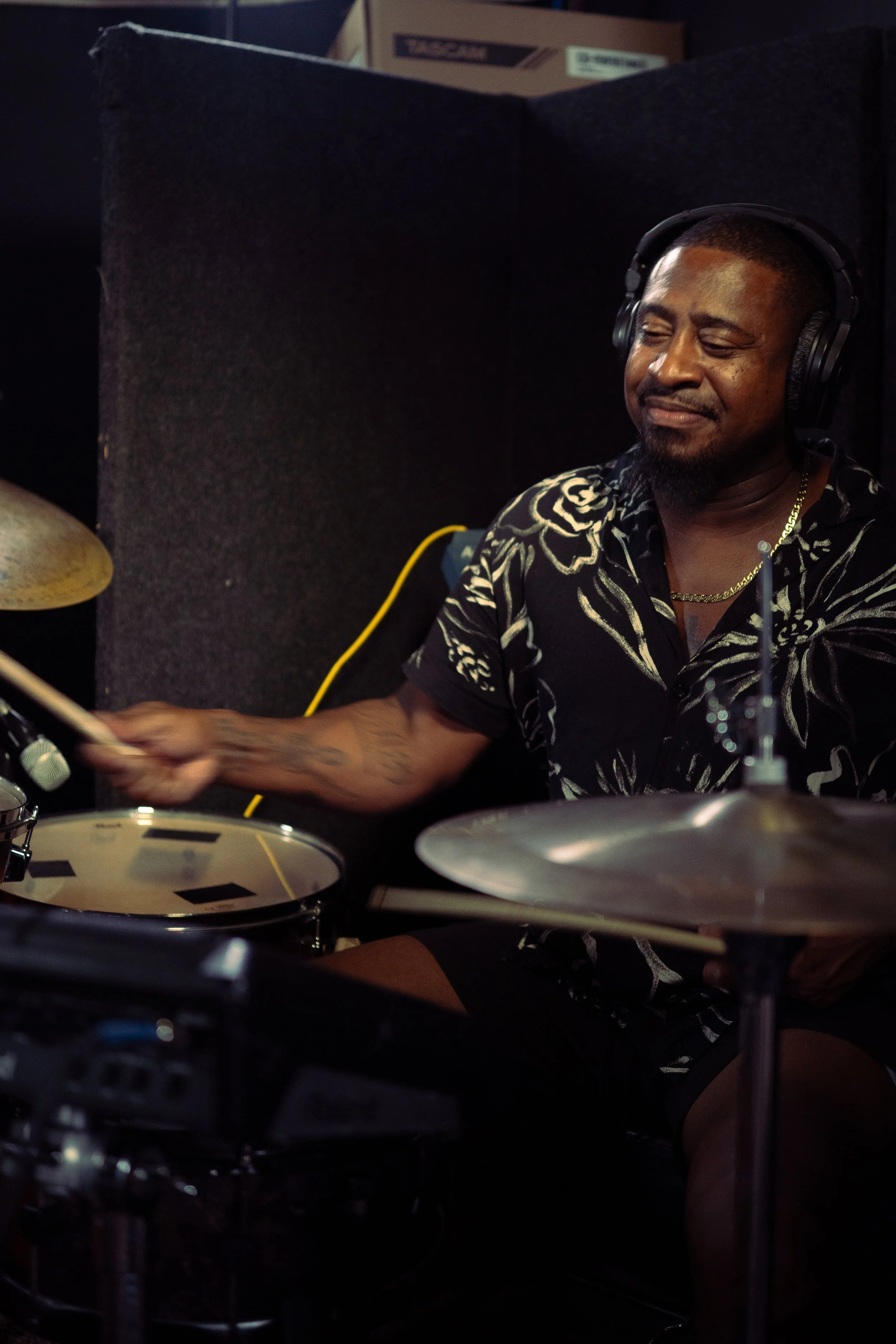 A man playing drums, wearing headphones and a black shirt with a floral pattern, in a music studio.