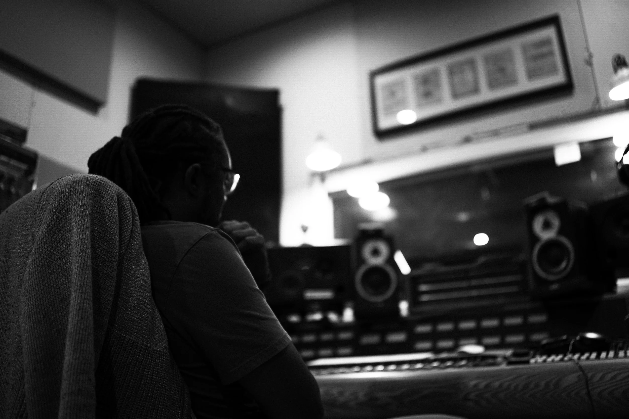 A person with dreadlocks, wearing glasses and a T-shirt, sitting in a music studio with sound equipment and monitors.
