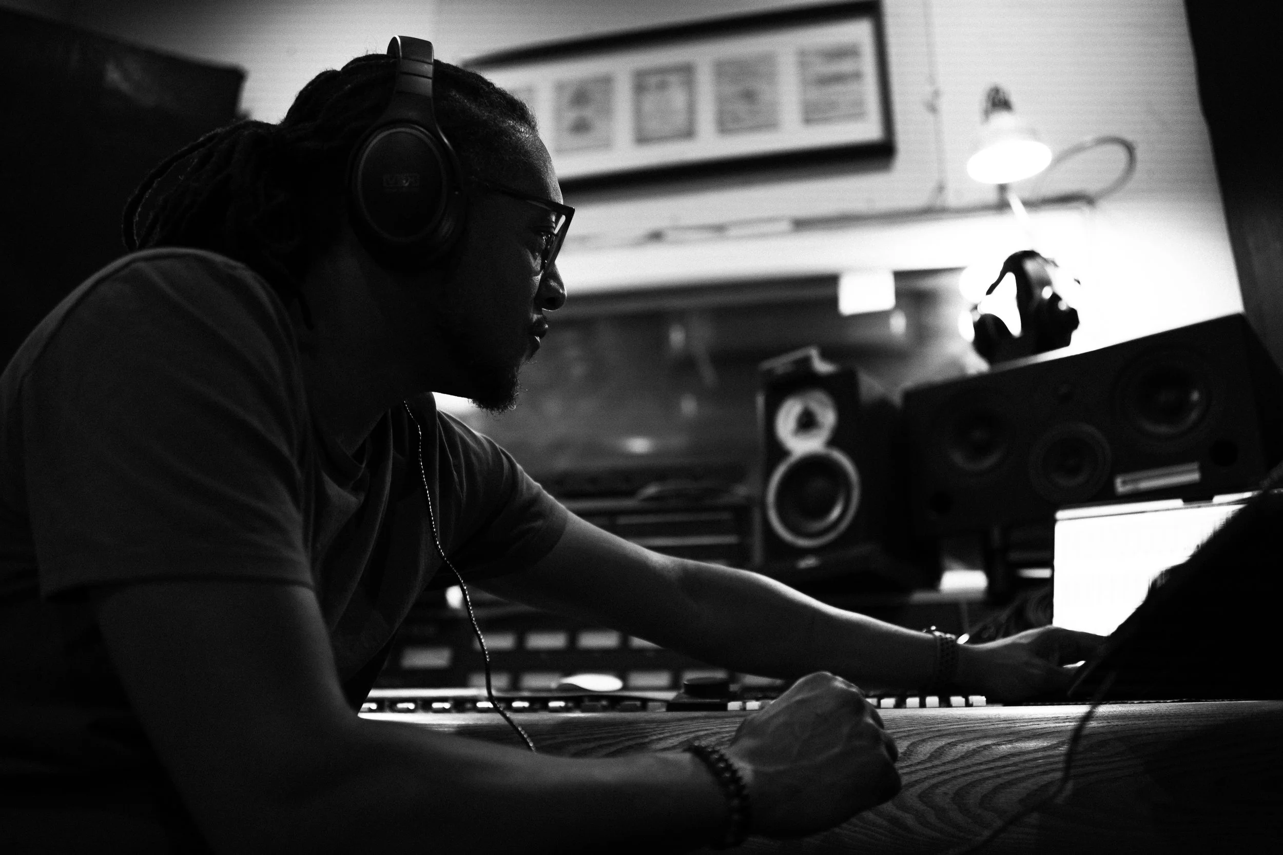 A man with dreadlocks and glasses working at a music production studio, wearing large headphones, with studio equipment and speakers in the background.