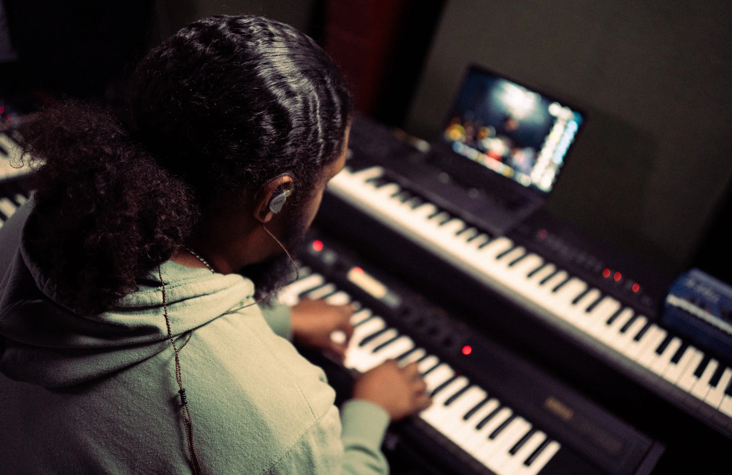 A person with curly hair wearing earbuds playing a digital keyboard with a computer setup in the background.