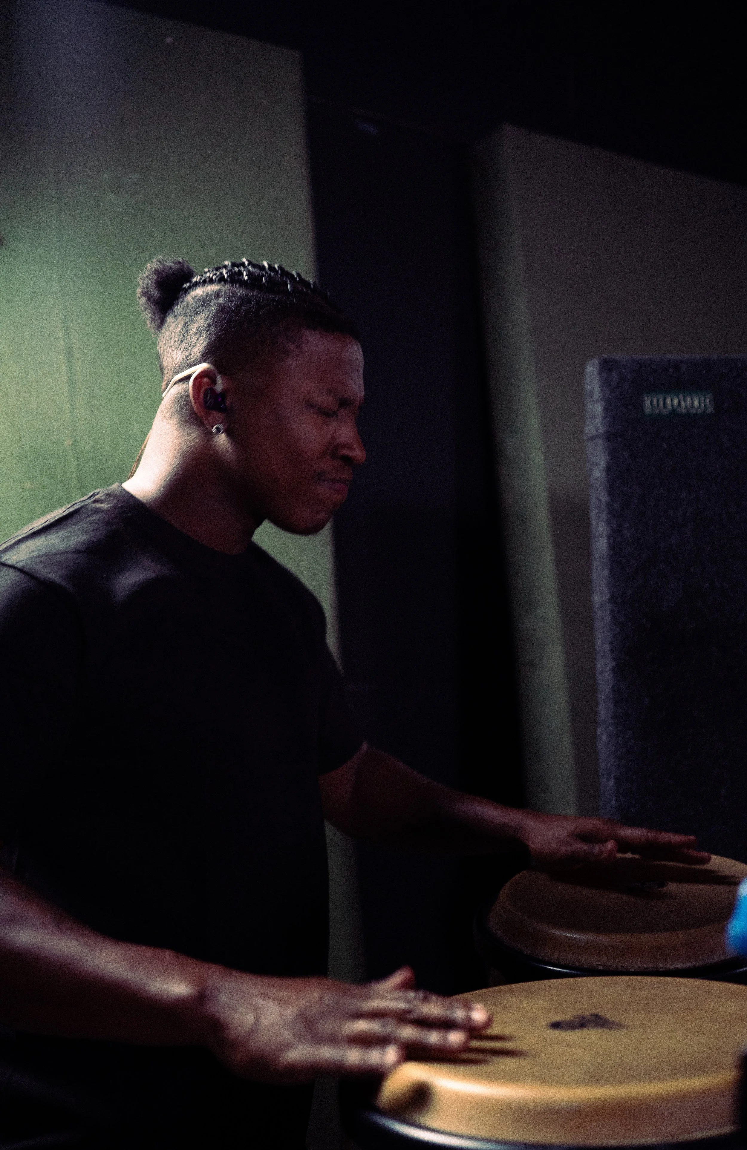 A young man with black hair in a bun, wearing in-ear monitors, playing percussion instruments with eyes closed in a dark room.