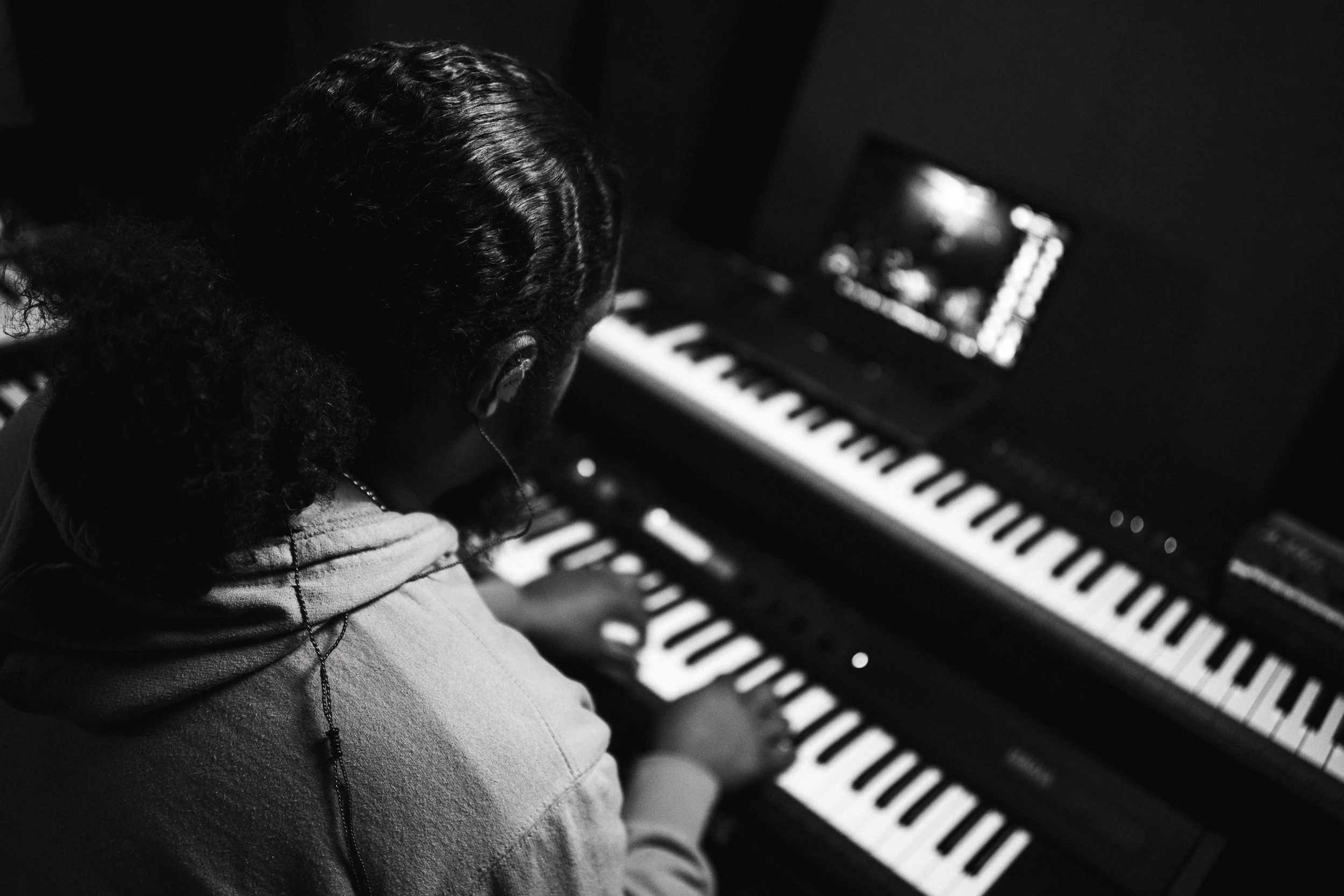 A person with long braids wearing a hoodie is playing a keyboard piano, viewed from behind in a dimly lit room.