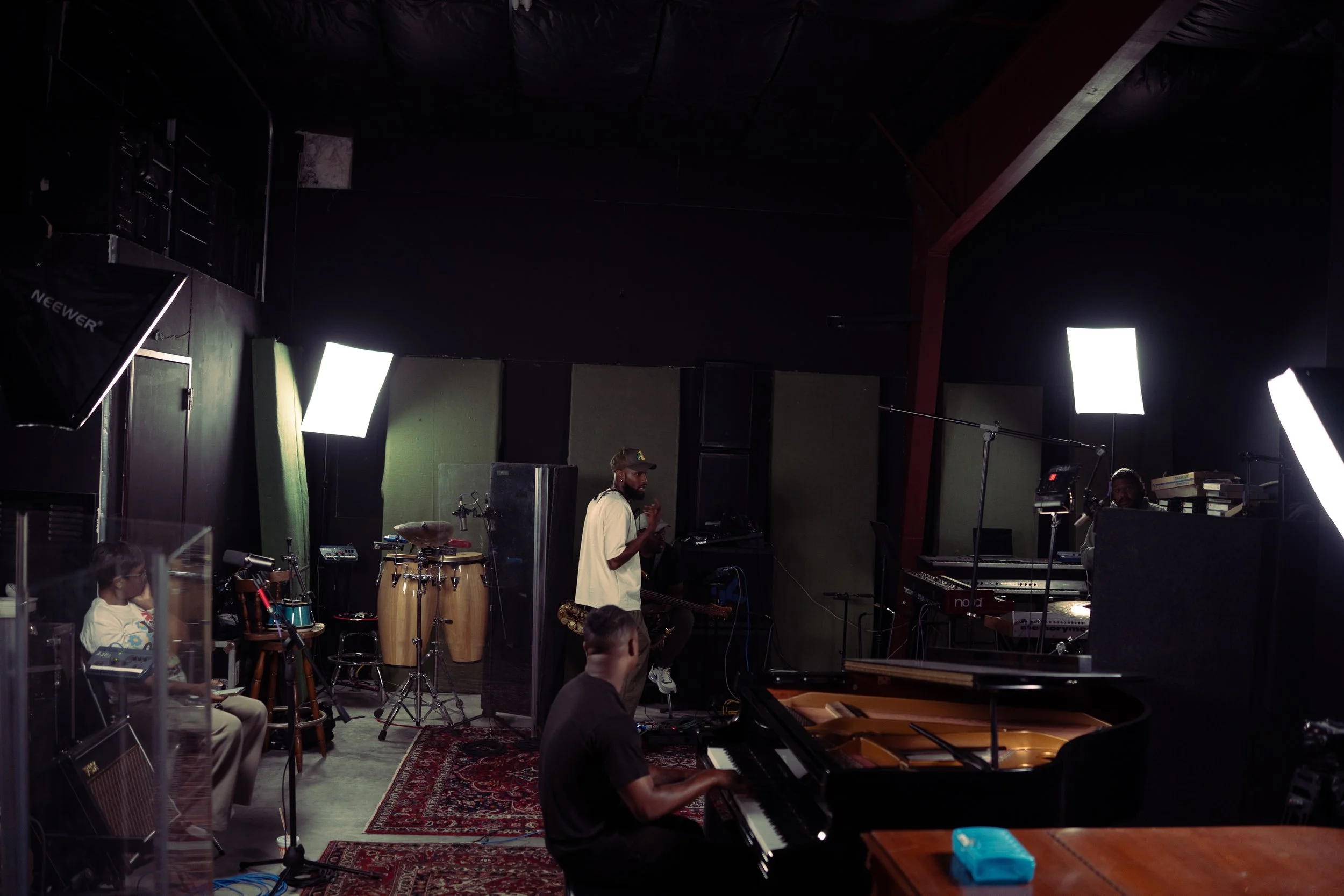 A music studio with a man playing a grand piano, a man sitting on a stool playing a keyboard, a person in a white shirt and a cap gesturing, and a person seated at a drum set. There are bright studio lights and music equipment in the room.