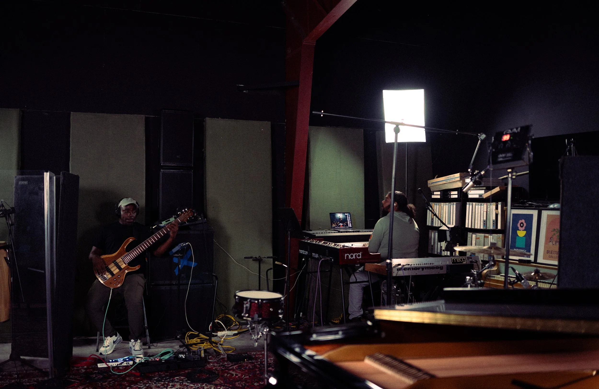Two musicians in a recording studio playing instruments, with one on bass guitar and the other on keyboard, surrounded by music equipment, books, and studio lighting.