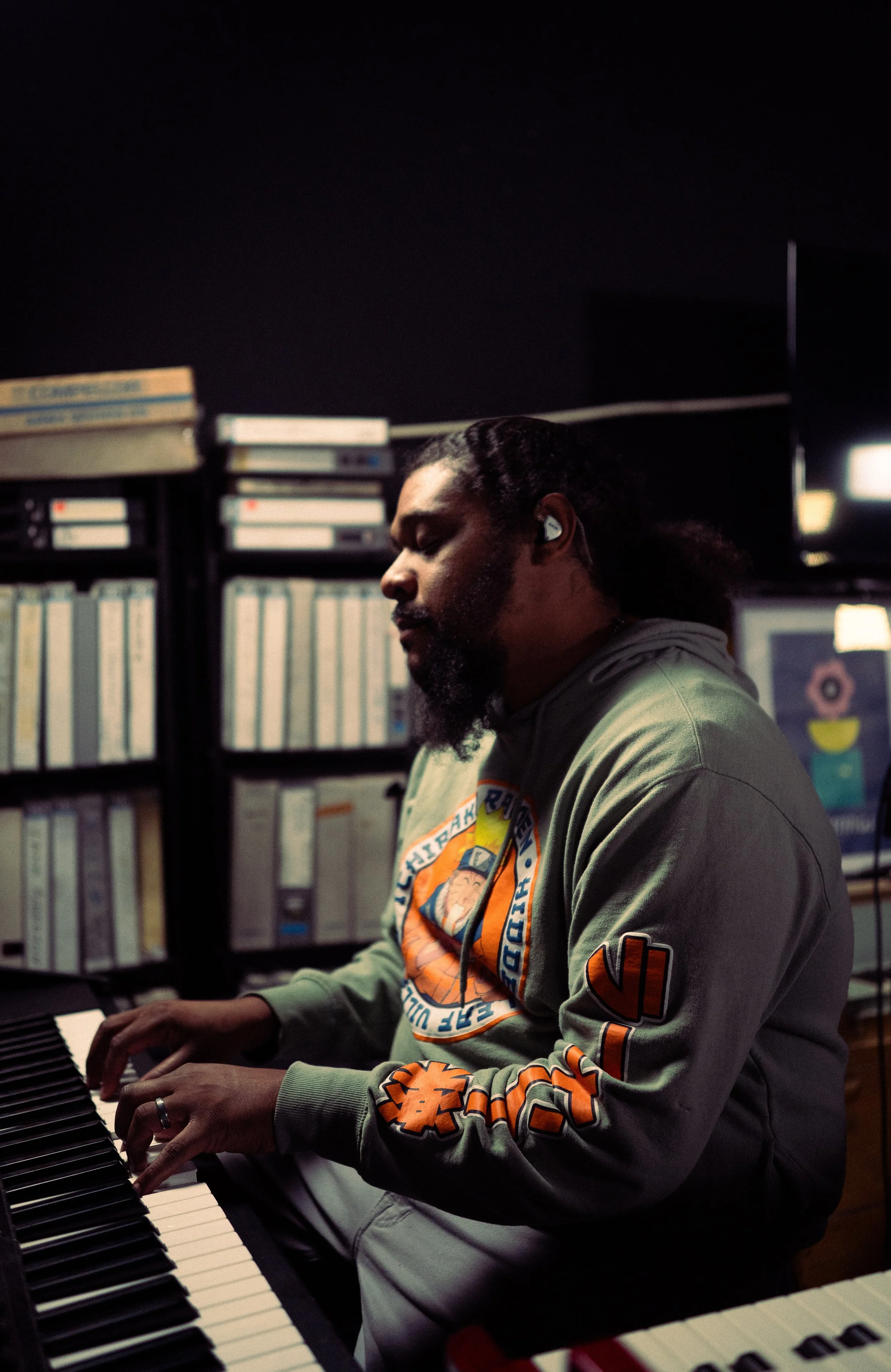 A man playing the piano in a dimly lit room, wearing gray headphones and a gray hoodie with colorful graphics and patches.
