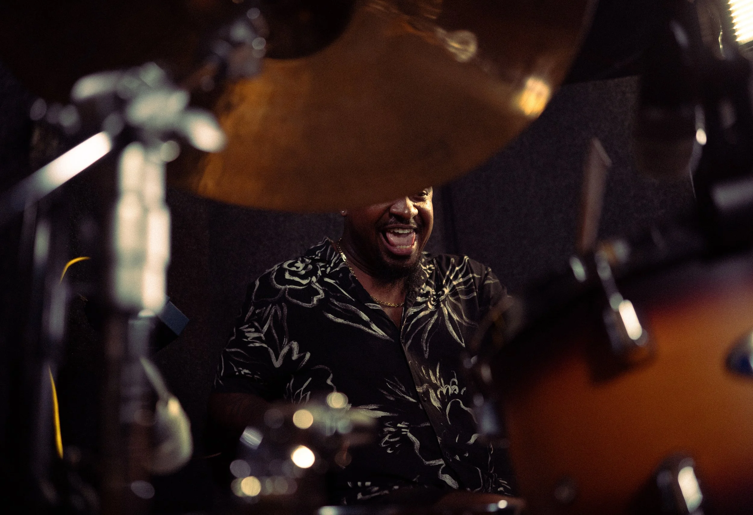 A man smiling and laughing while playing the drums, seen through the drum set.