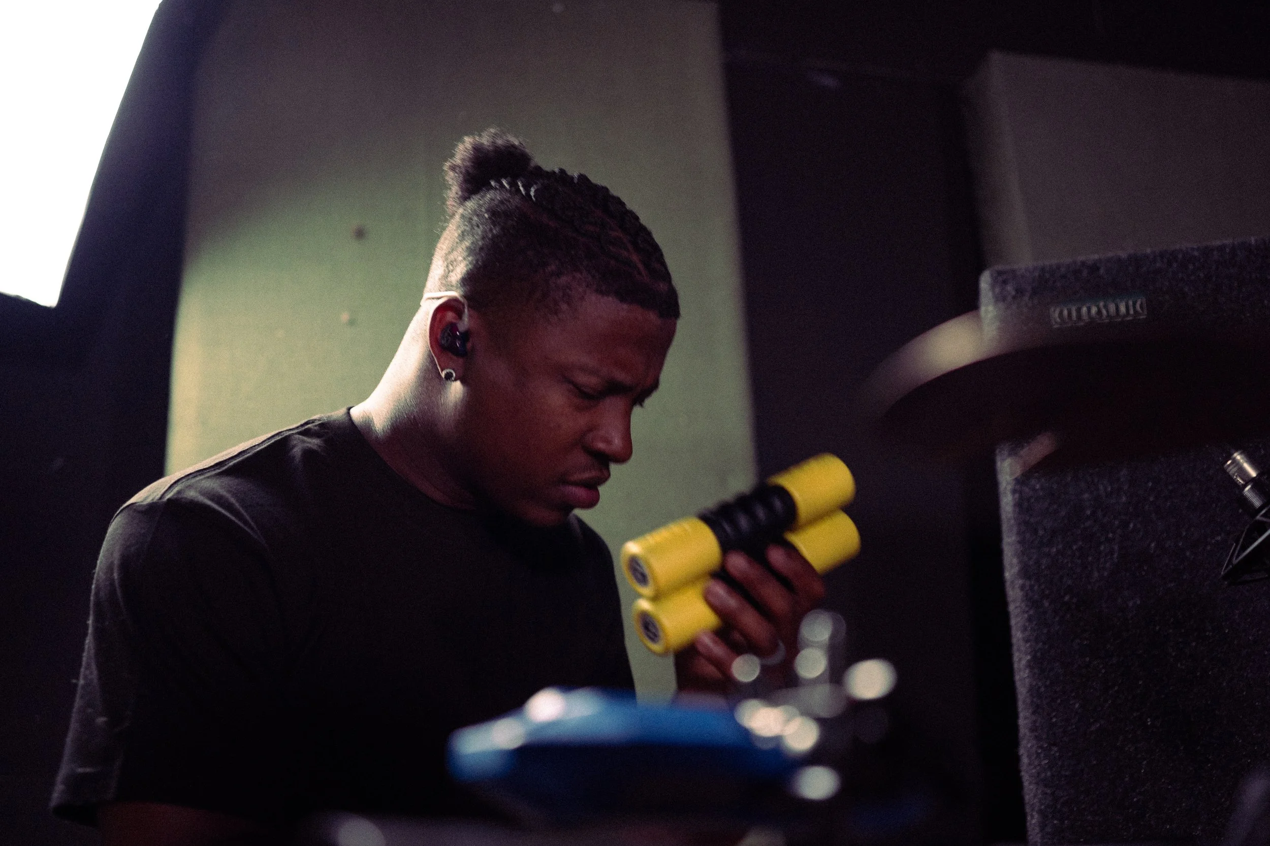 A person with braided hair and earrings holding yellow foam rollers looks downward in a dimly lit room, possibly a recording studio.