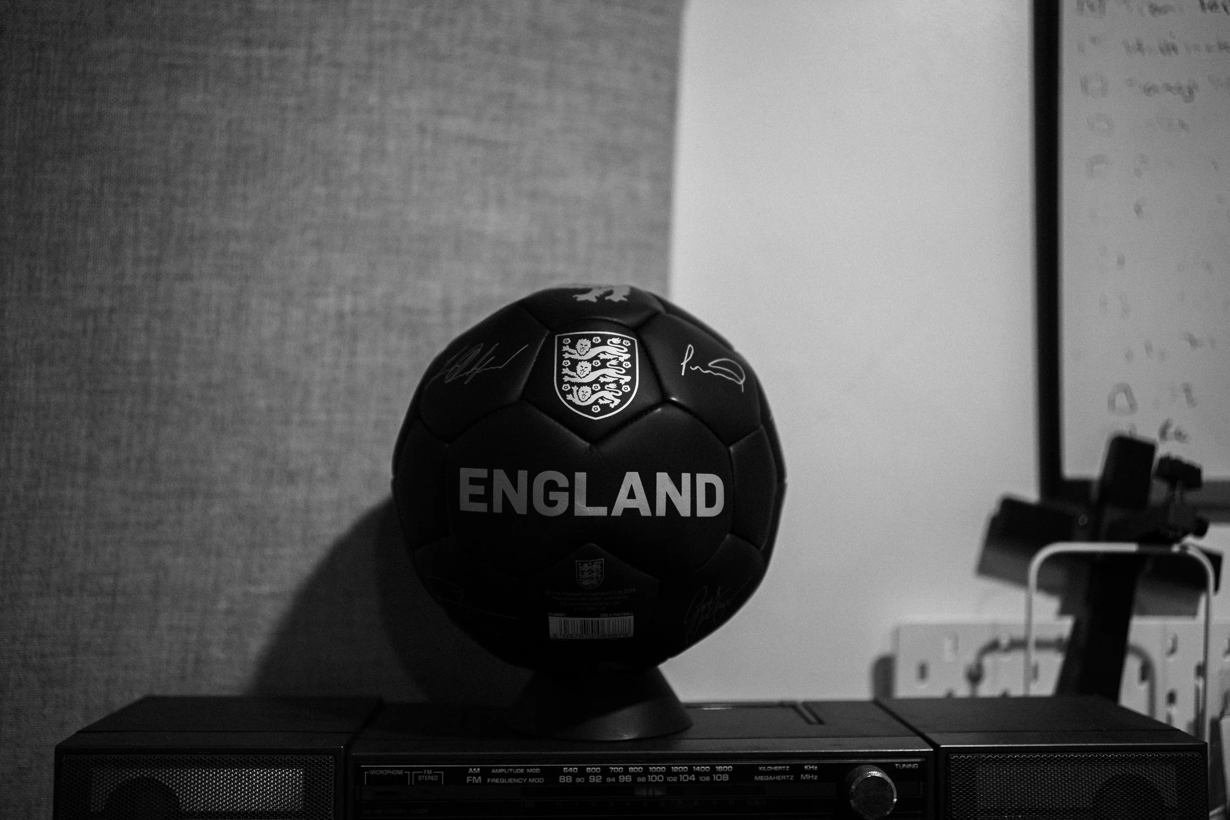 Black football with England's emblem and signatures on it, placed on a stand above a vintage radio, with a whiteboard and electrical outlets in the background.