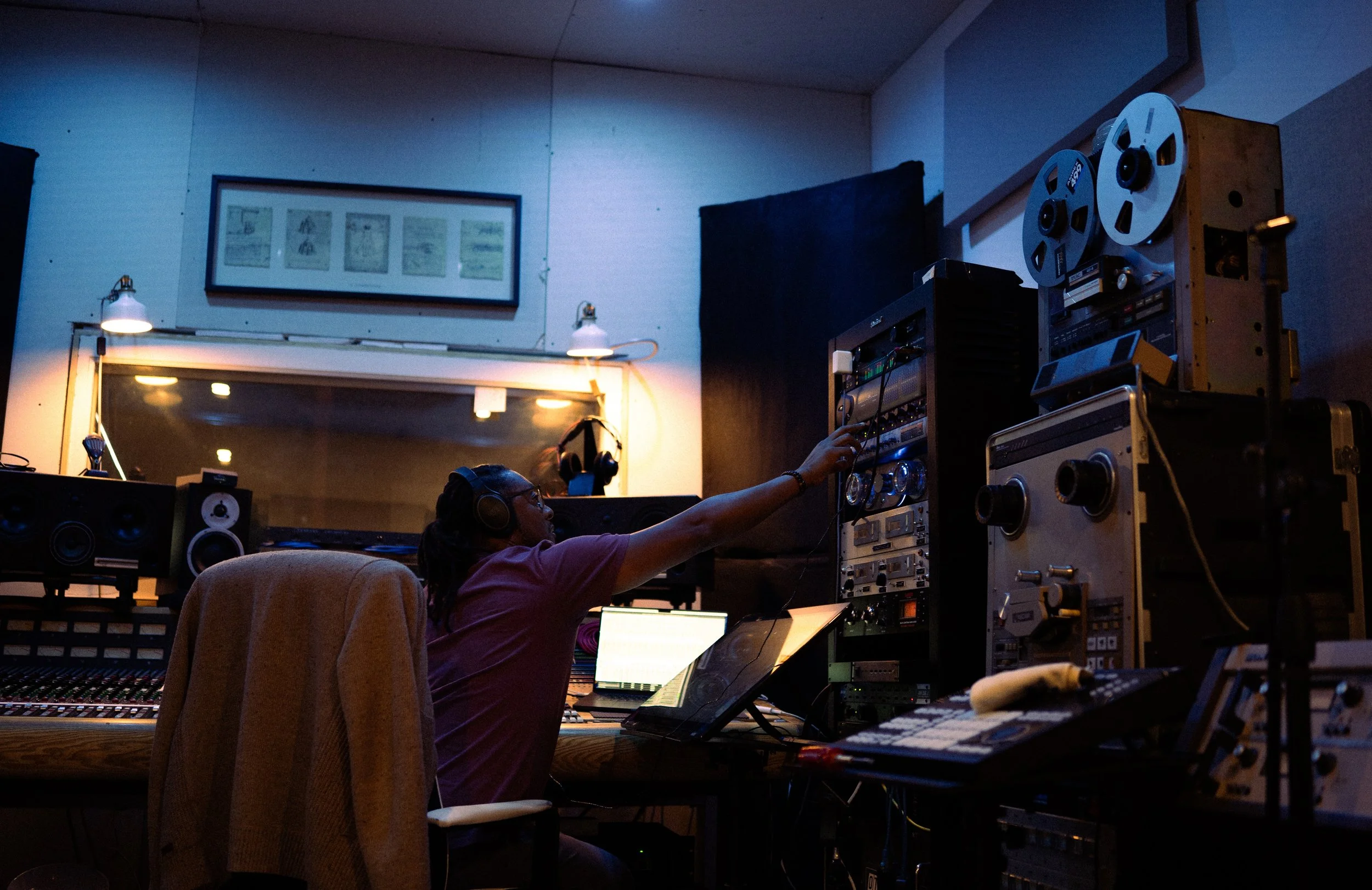 A person with headphones working in a recording studio, adjusting audio equipment amidst various electronic gear and sound mixing consoles.