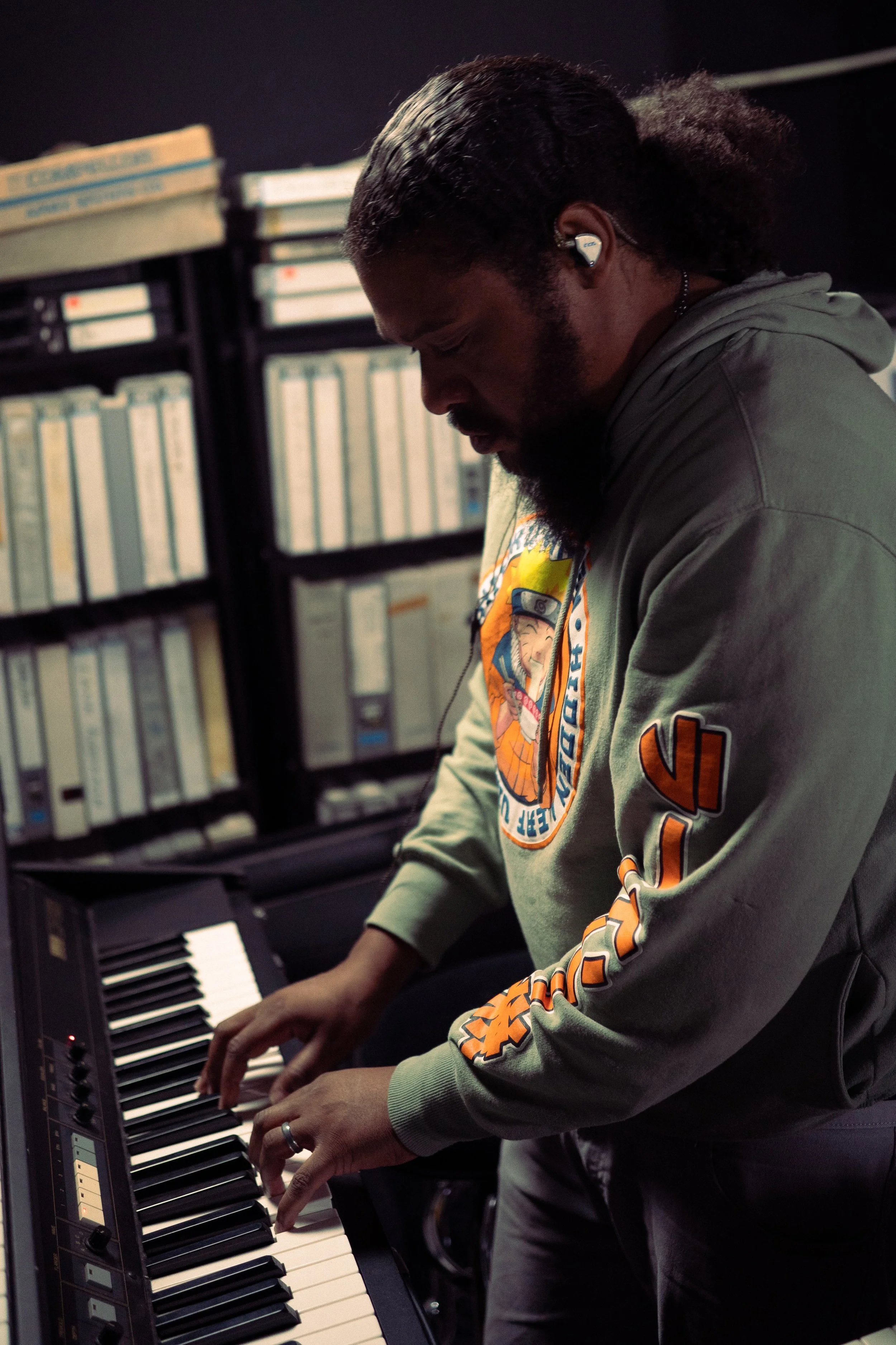 A man with a beard and long hair tied back playing a keyboard in a room with shelves of binders behind him. He is wearing a hoodie with Naruto and Dragon Ball Z patches and earbuds.