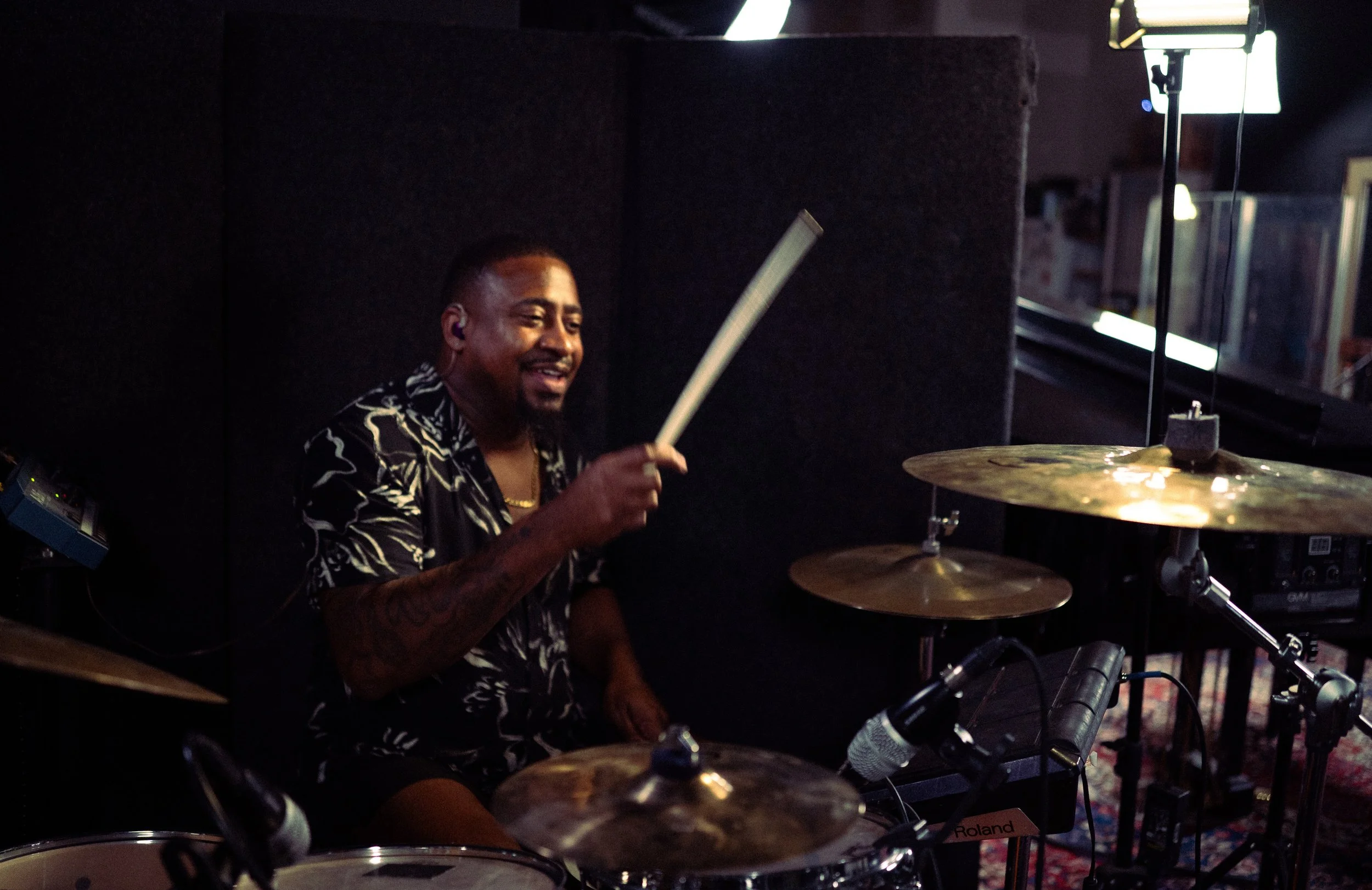 A man playing the drums in a recording studio or music space, smiling and holding a drumstick.