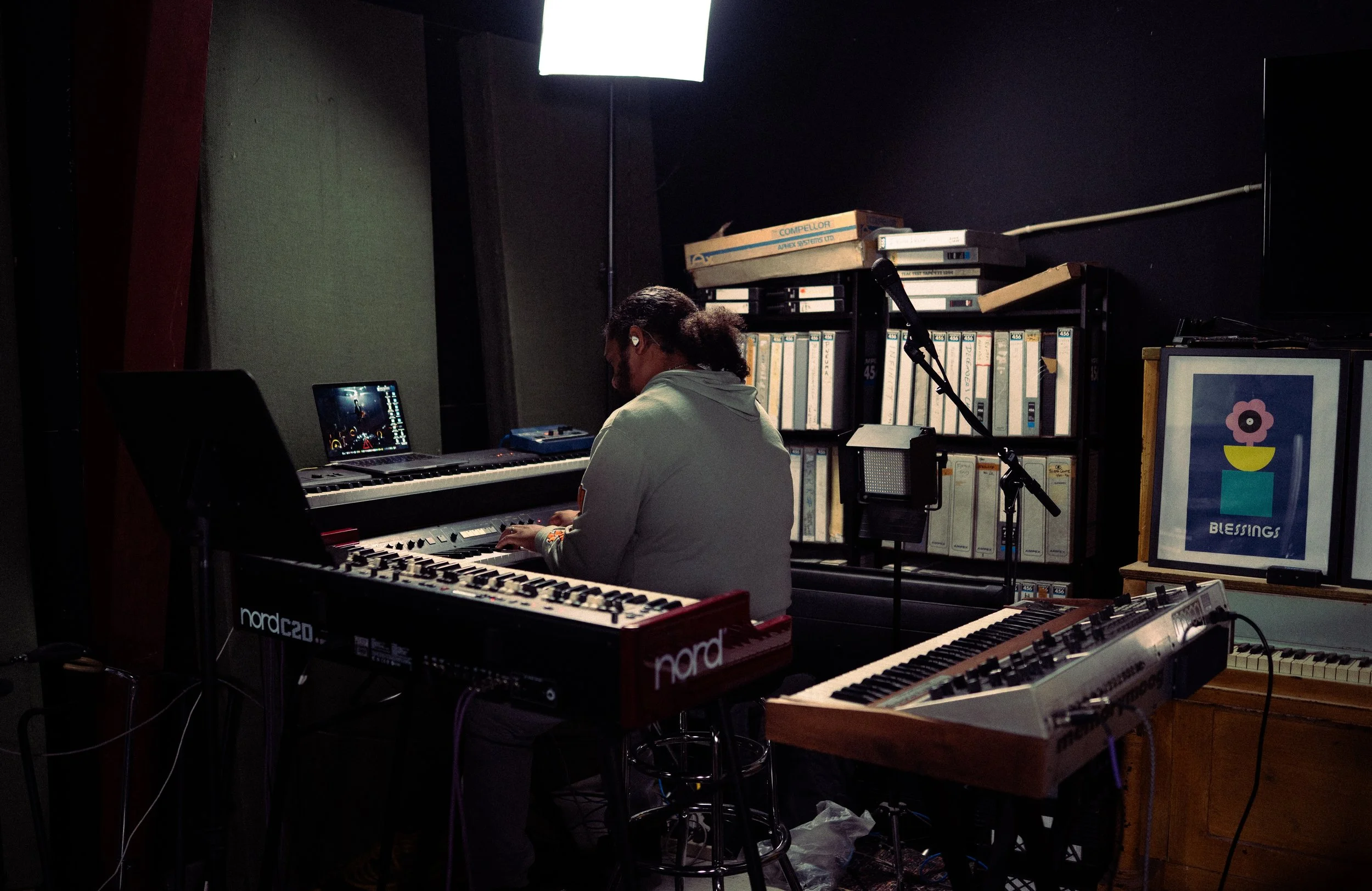 A person sitting at an electronic keyboard in a recording studio surrounded by music equipment and shelving with binders, with studio lighting and a framed picture that says 'Blessings' on the wall.