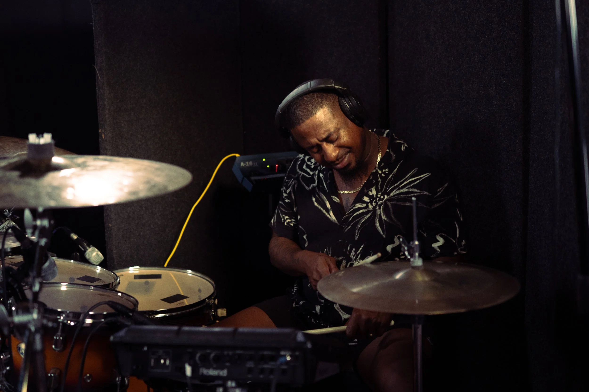 A man playing a drum set in a music studio, wearing headphones and a black shirt with white leaf patterns.