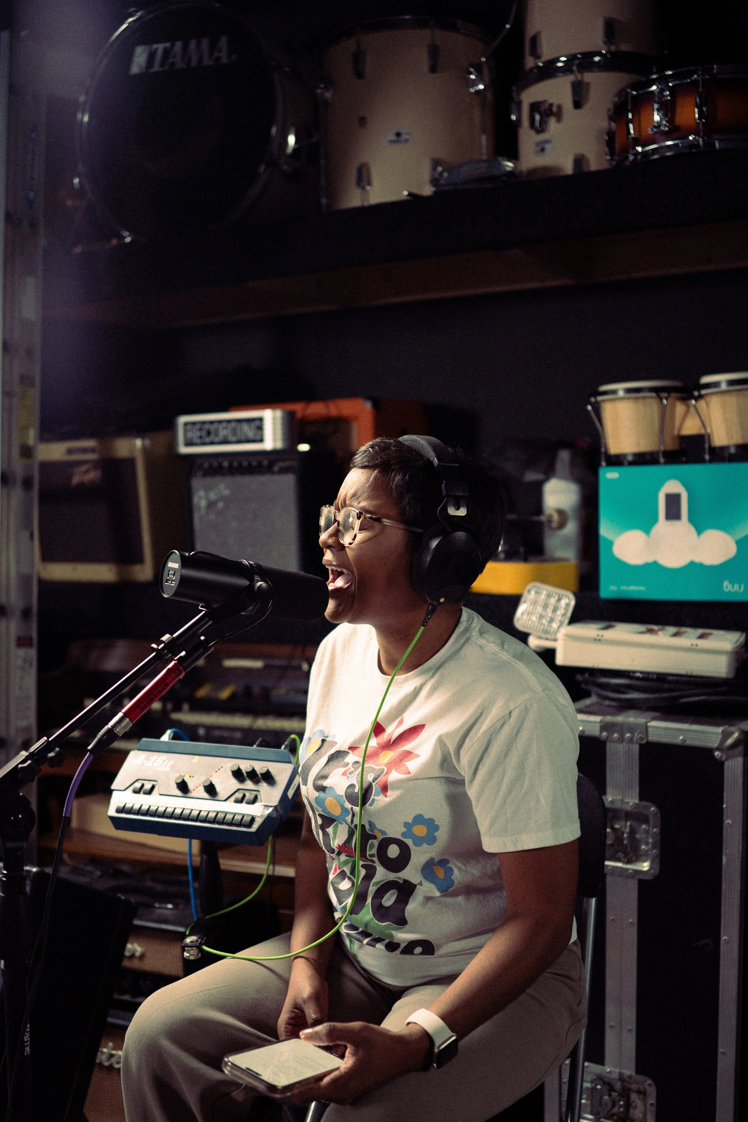 A woman wearing glasses and headphones singing into a microphone in a music studio. She is sitting on a chair, holding a smartphone in her left hand. The background shows musical equipment, including drums and amplifiers.
