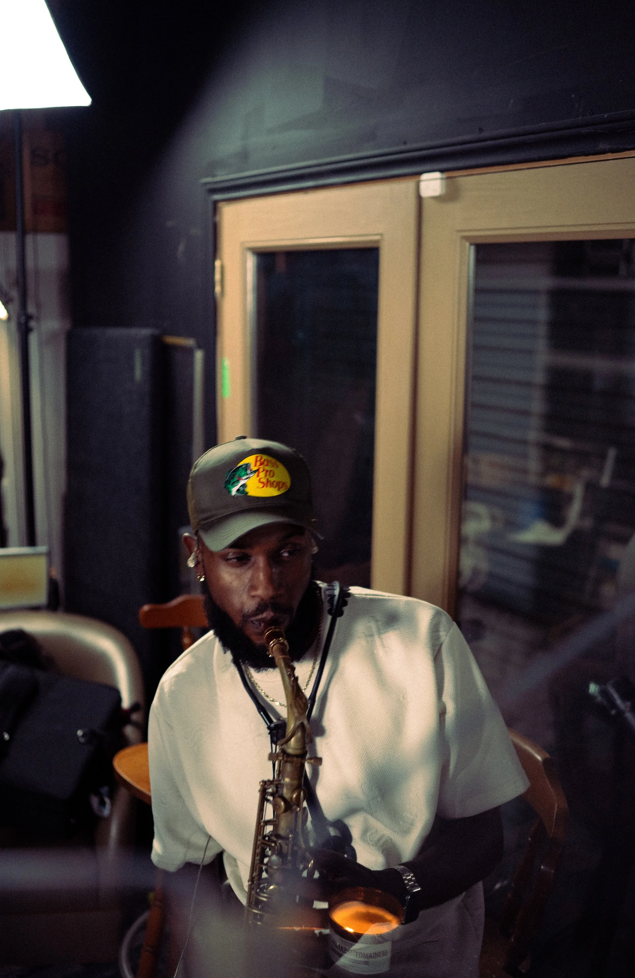 A man wearing a bass pro shop hat and white shirt playing a saxophone in a dimly lit indoor setting.