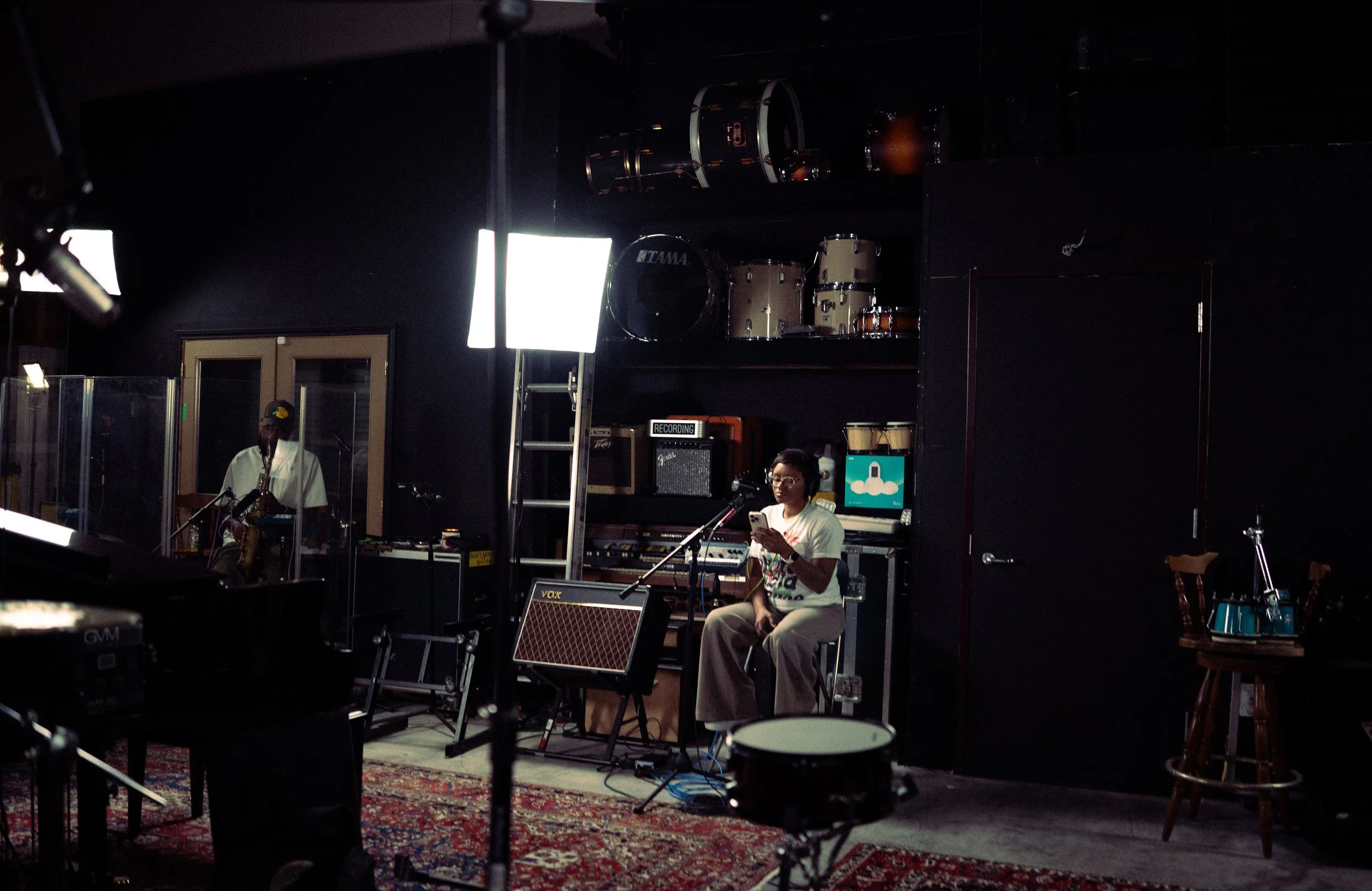 A woman sitting on a stool in a recording studio, looking at her phone, with a microphone in front of her. A man is playing the saxophone behind a clear partition, with musical instruments and recording equipment around.